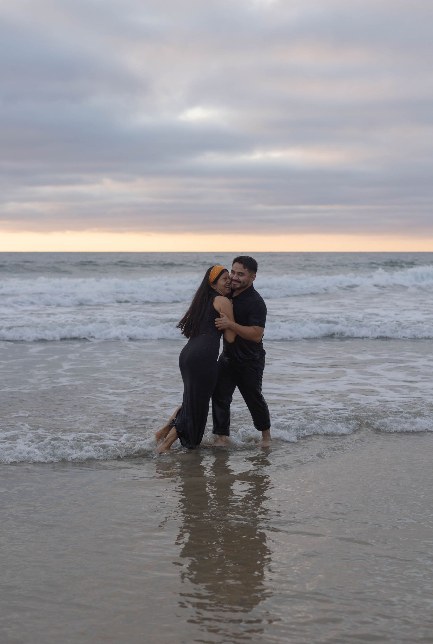 Couple embracing and kissing on a sandy beach at sunset, standing in shallow ocean waves wearing black outfits. La Jolla Shores, San Diego.