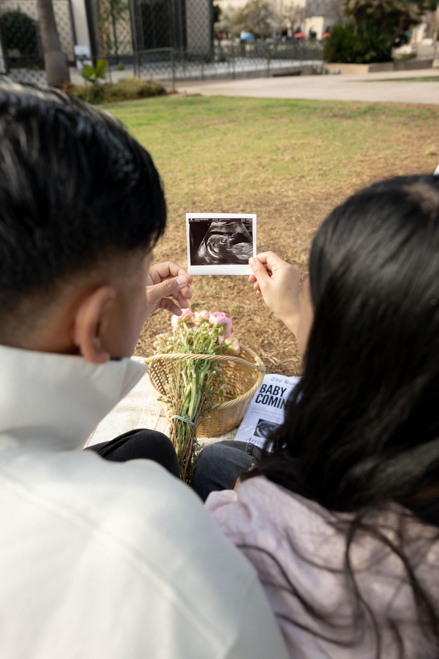 Couple holding ultrasound photo and flowers outdoors, pregnancy announcement with baby coming sign. Family photographer San Diego California