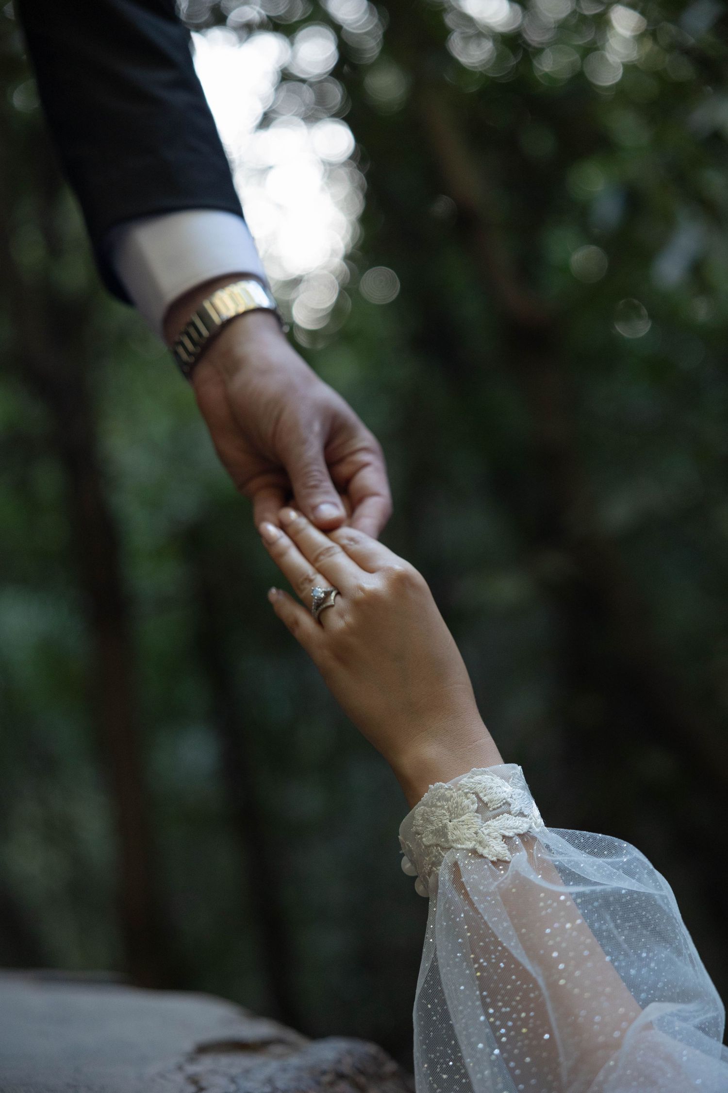 Bride and groom hold hands gently in a lush forest, bride wearing lace sleeve gloves and diamond ring.