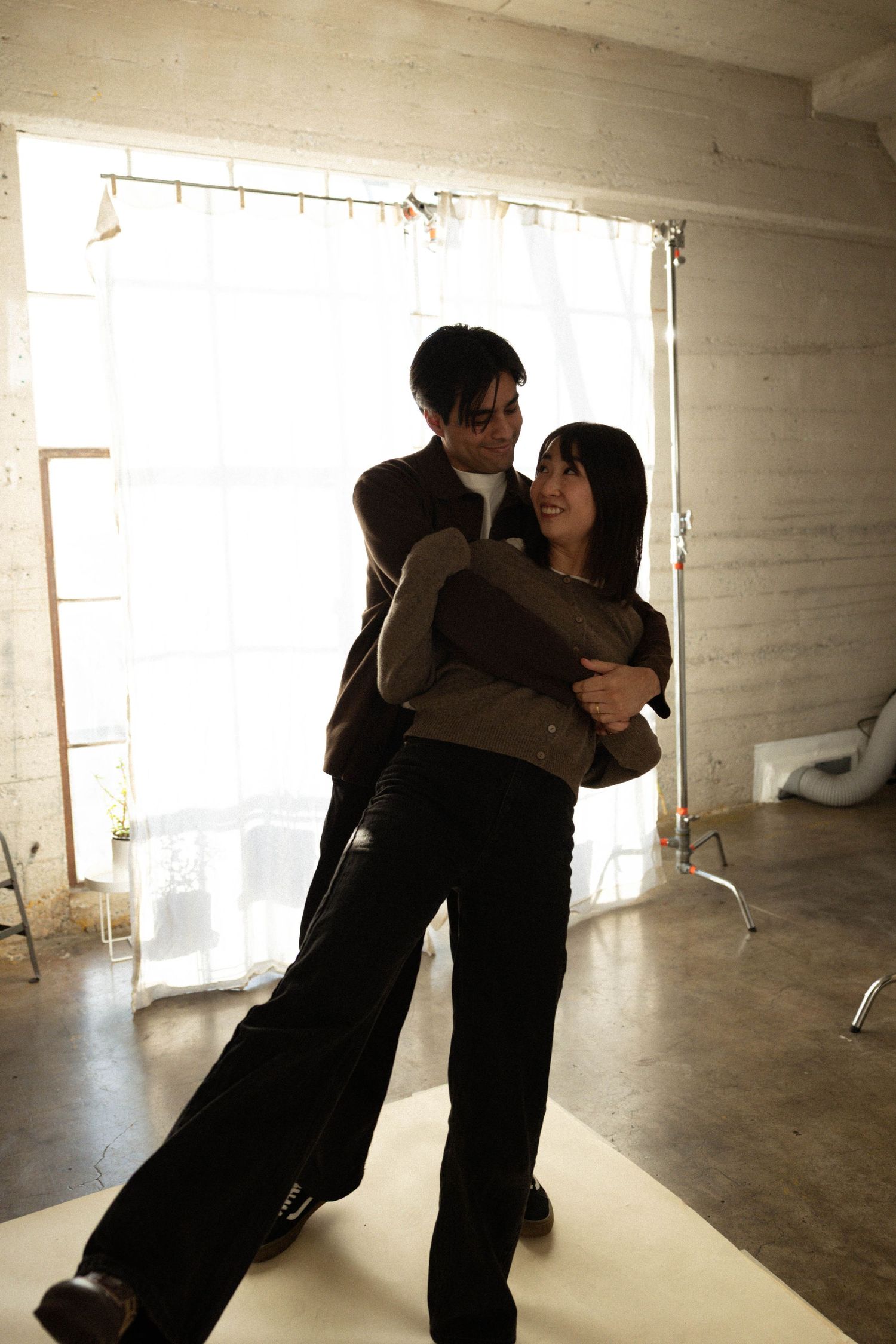 A couple embraces and dances together in a bright studio space with white curtains and concrete walls. Couples photographer.