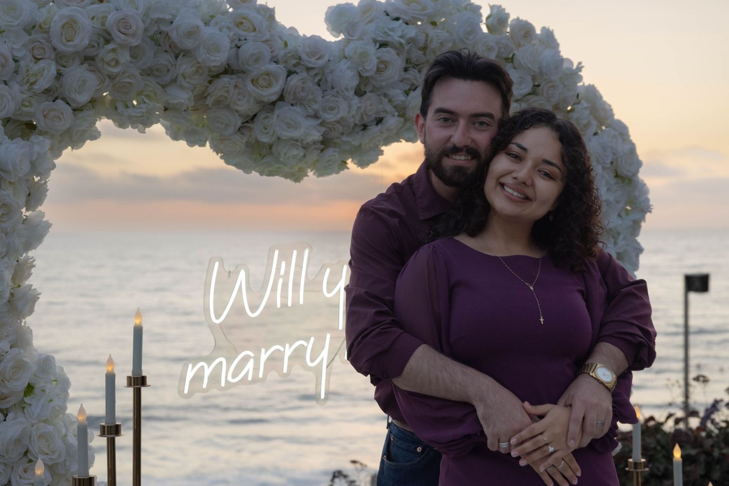 Couple embracing under white floral arch at sunset with Will You Marry Me text overlay by the ocean. Portrait Photography.