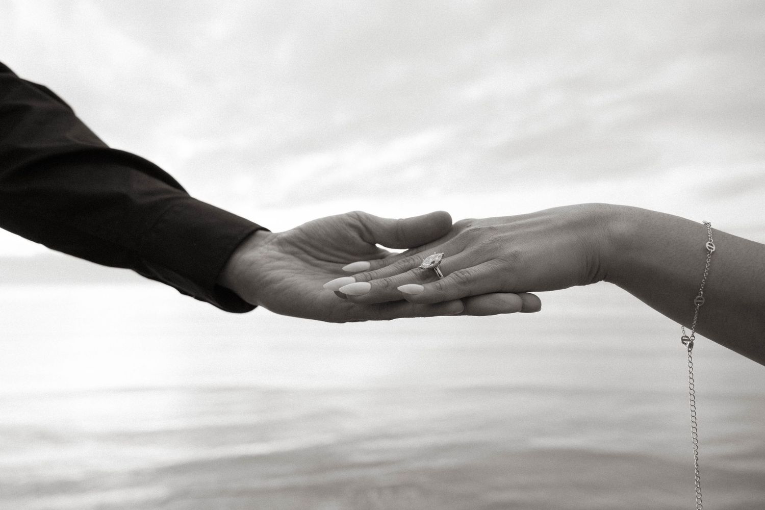 Black and white photo of two hands touching, woman wearing engagement ring near ocean backdrop.