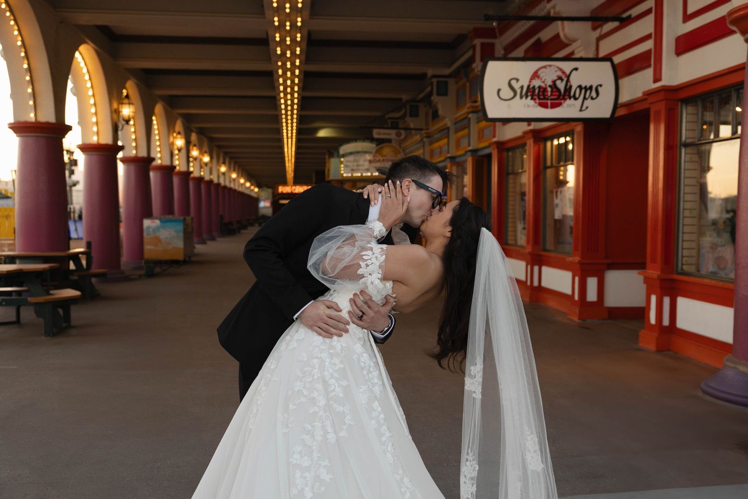 Bride and groom sharing a romantic dip kiss at an amusement park arcade, bride in white gown with veil.