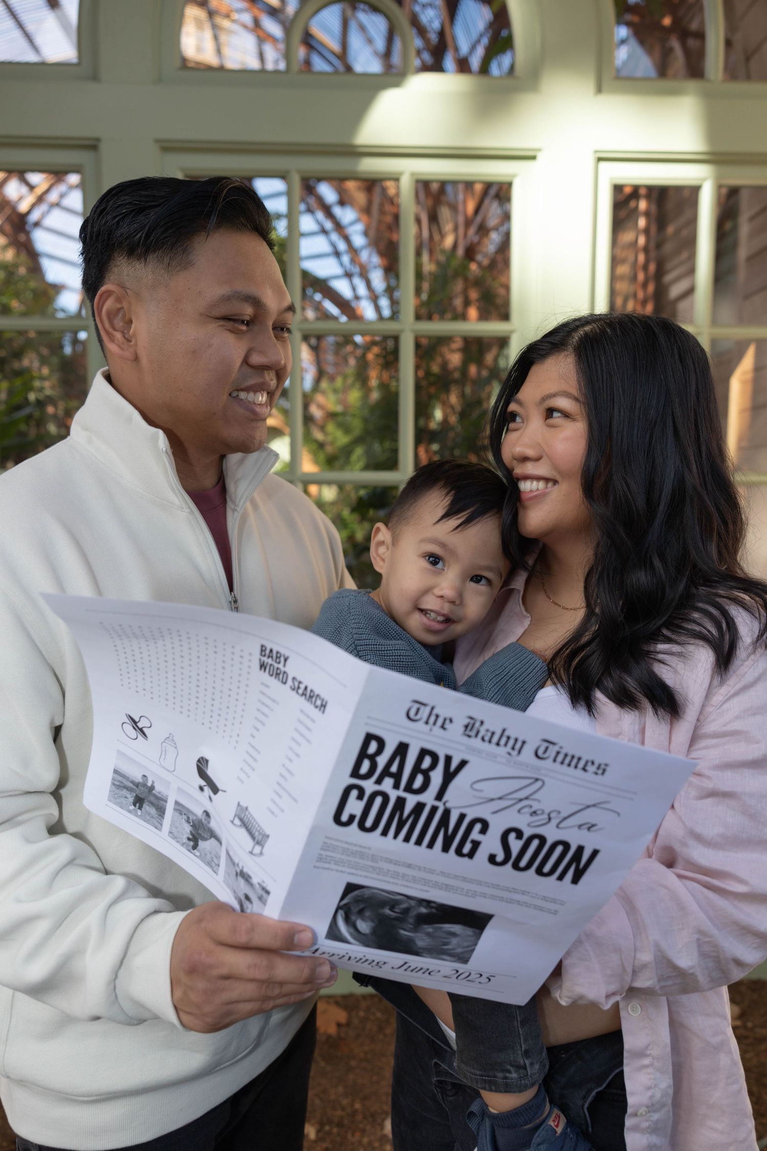 Happy family holding a Baby Coming Soon newspaper announcement with toddler son outdoors.