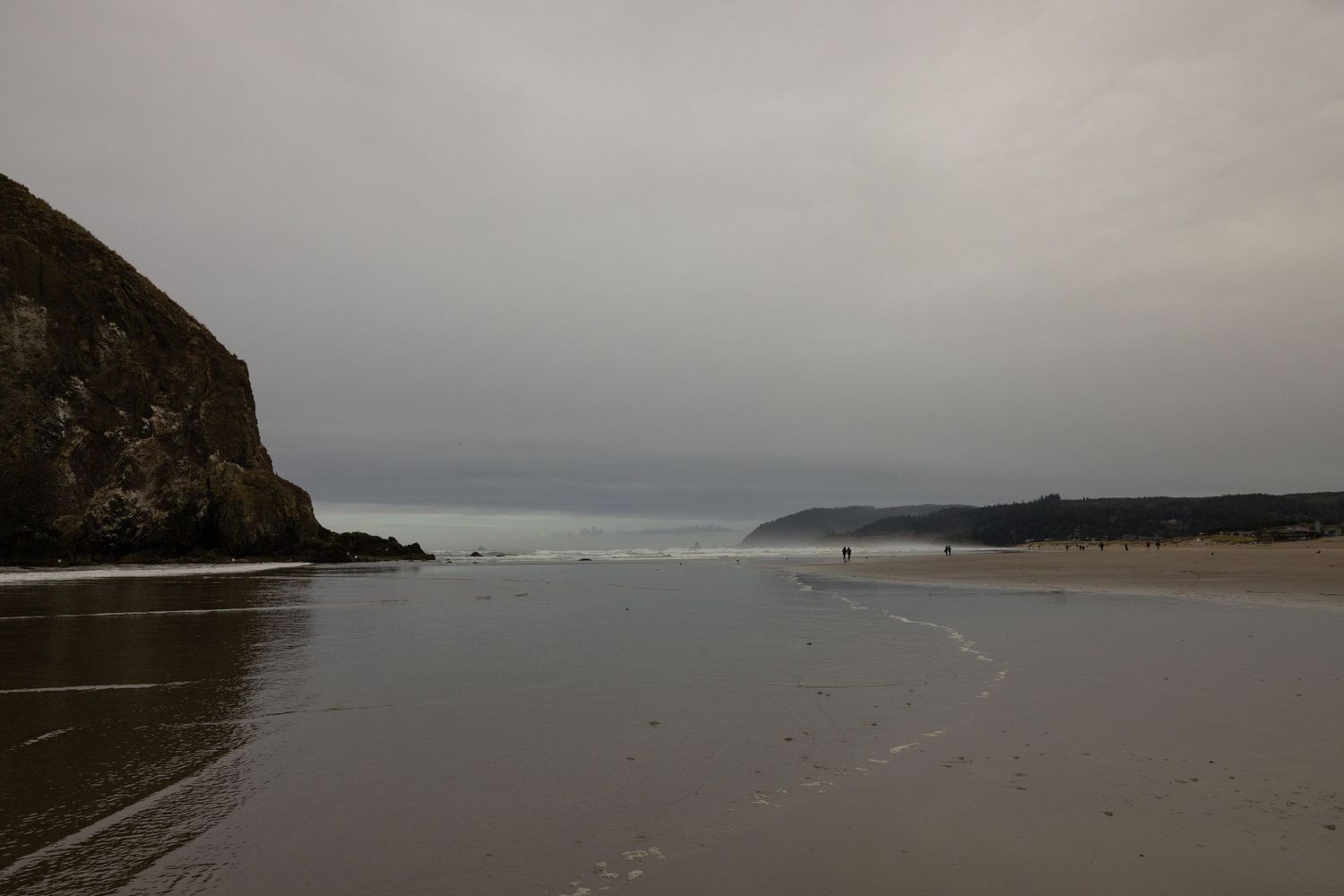 A moody overcast beach with wet sand, dramatic cliffs, and distant figures along the shoreline.