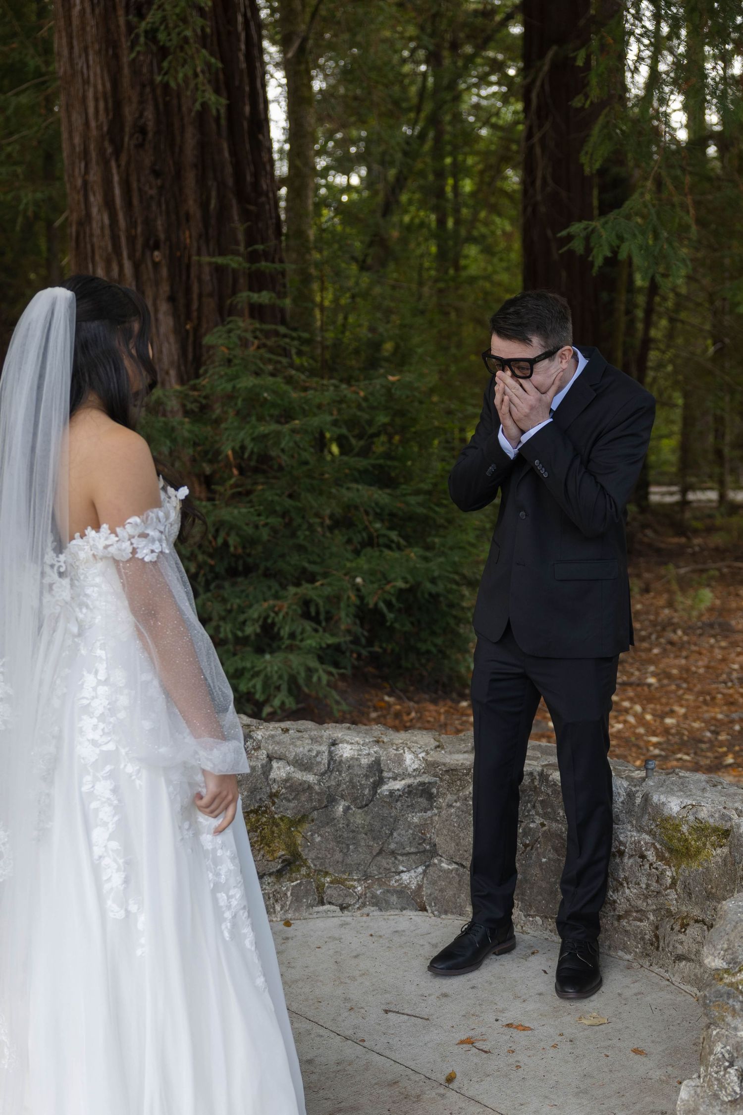 Groom covers his mouth in emotional reaction seeing bride in white lace wedding gown during forest first look ceremony. Elopement Photos.