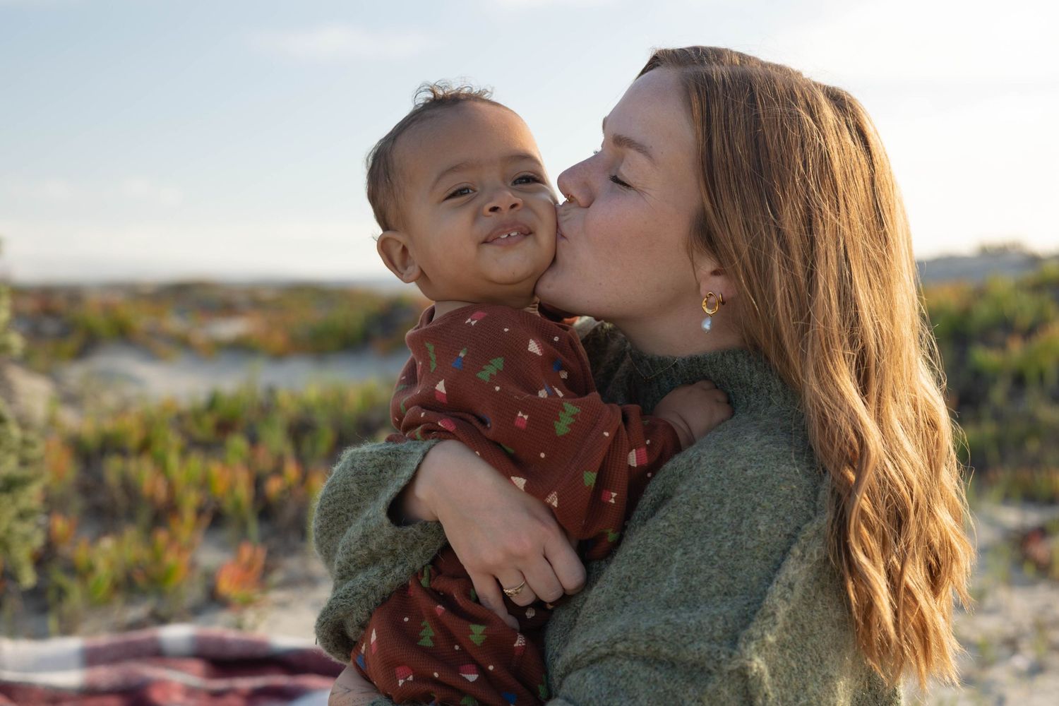 A mother kisses her smiling baby on the cheek at a sunny beach. Family Photographer.