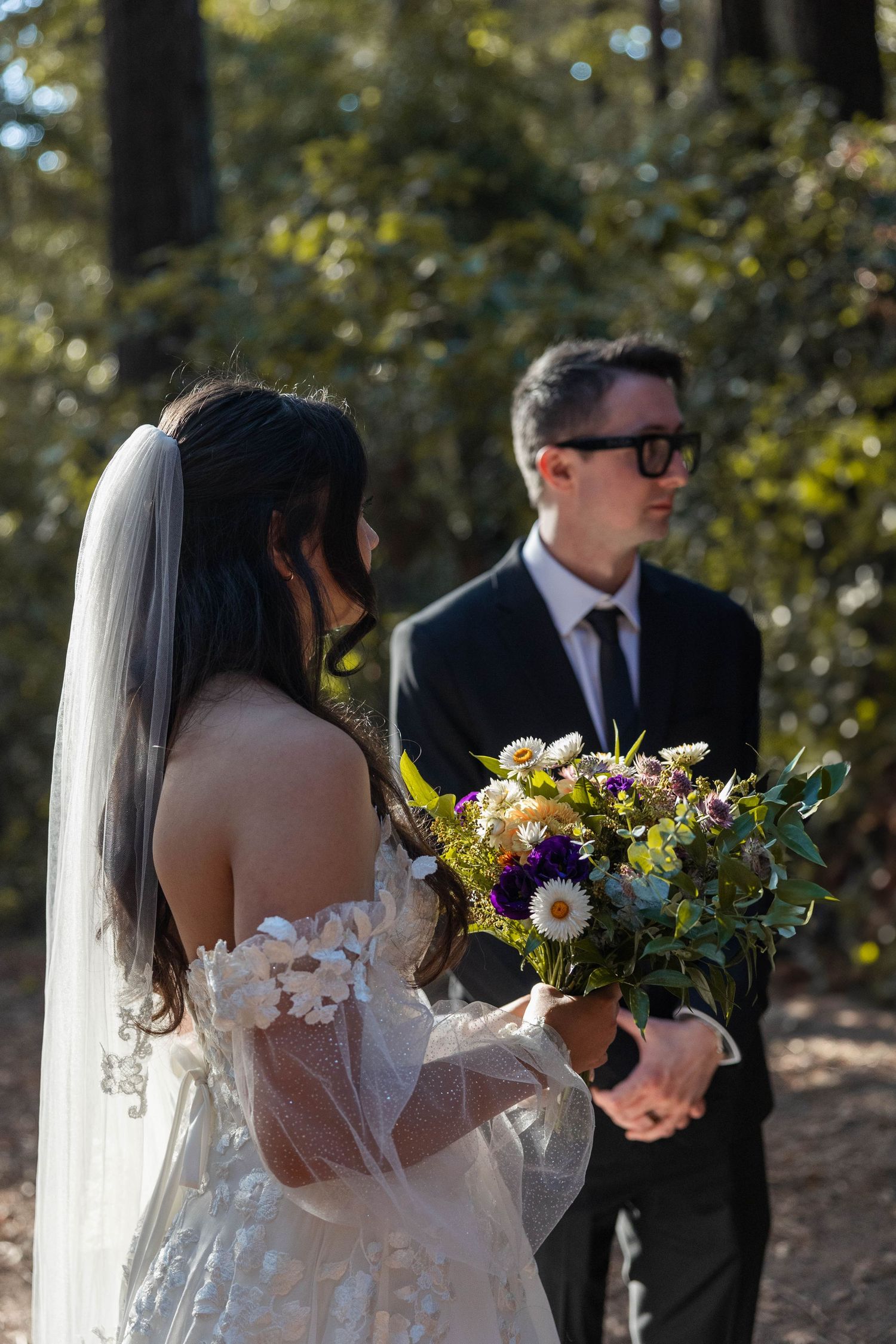 Bride in lace off-shoulder gown holding wildflower bouquet stands before groom in black suit during forest wedding ceremony. Elopement Photo