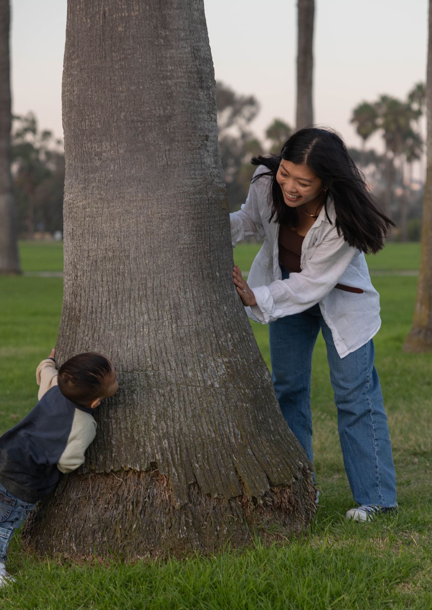 A woman and child playing hide and seek around a large tree trunk in a green park. Family Photographer.