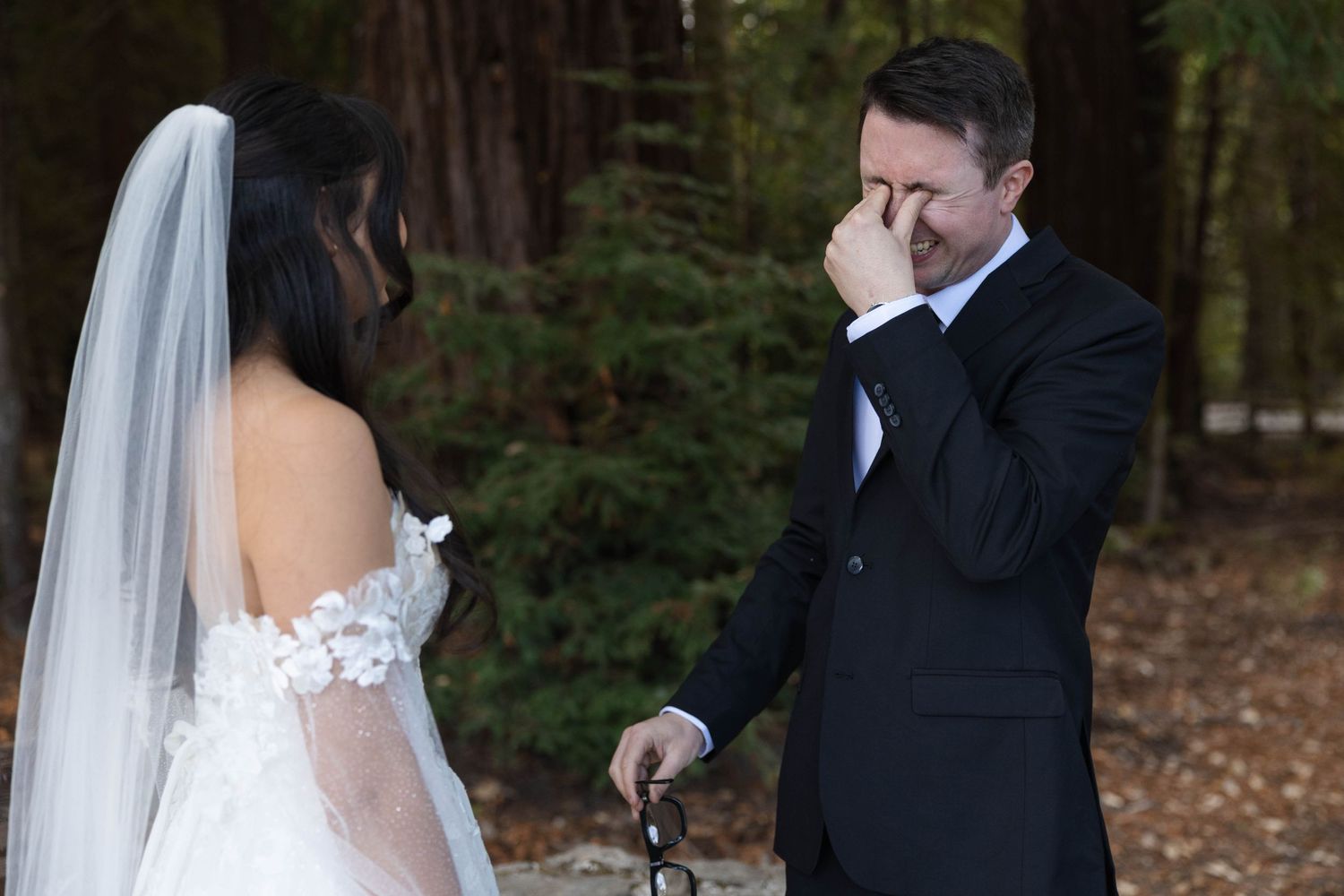 Emotional groom wipes tears seeing bride for first time in sunlit redwood forest.