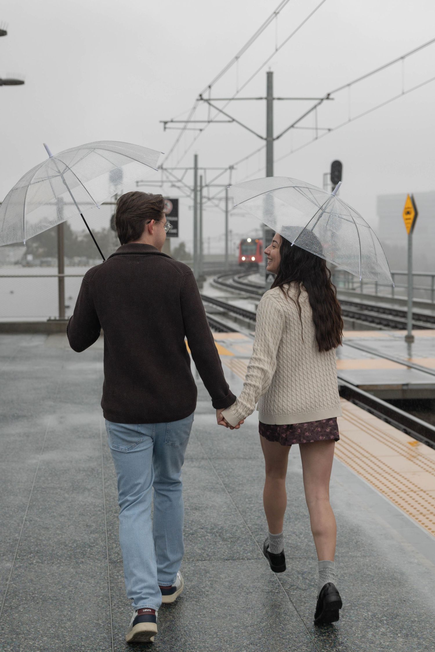 couple holding hands and walking away from camera in the rain holding transparent umbrellas.