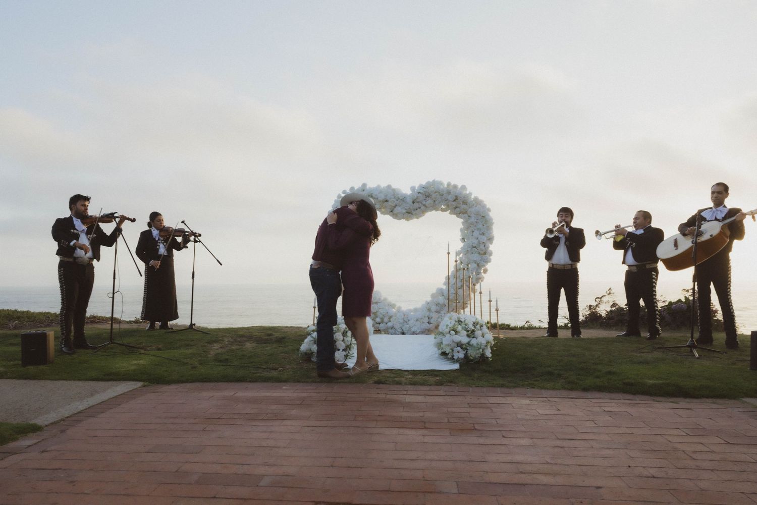 Couple embracing under heart-shaped floral arch on wooden deck with mariachi band performing at outdoor wedding ceremony.