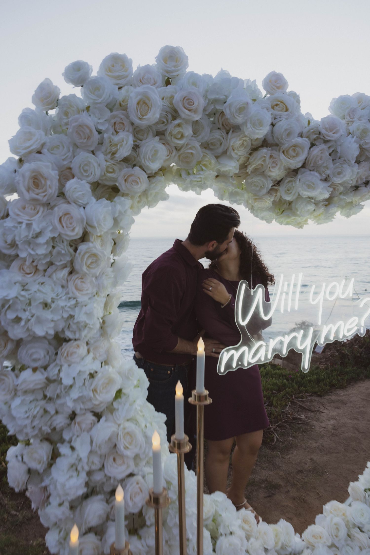 Couple kissing under a white rose floral arch with candles and a Will You Marry Me neon sign by the ocean. Portrait Photography.