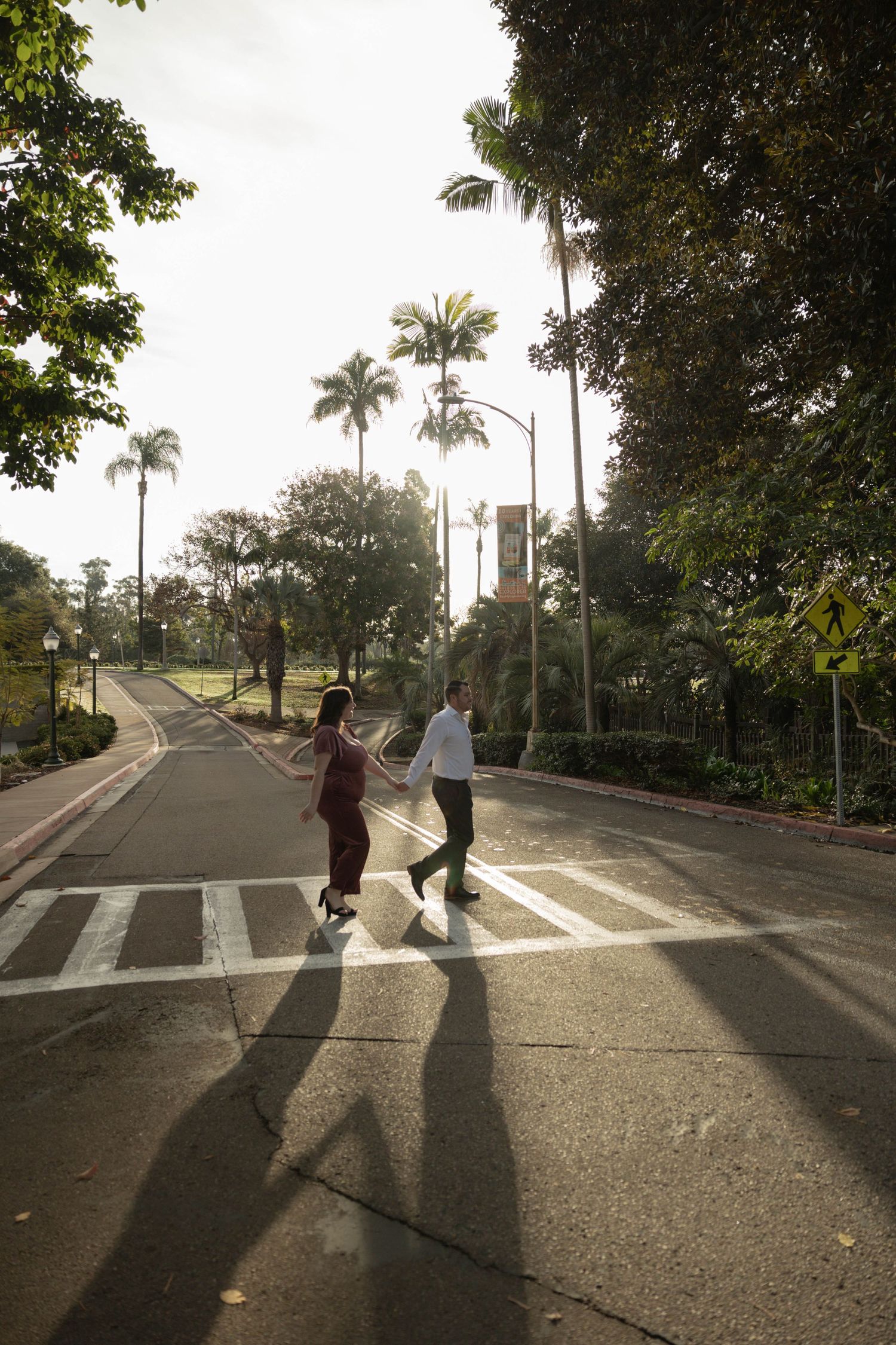 Couple holding hands crossing a zebra crossing on a tree-lined road with palm trees and warm backlight. Couples Portraits San Diego Ca