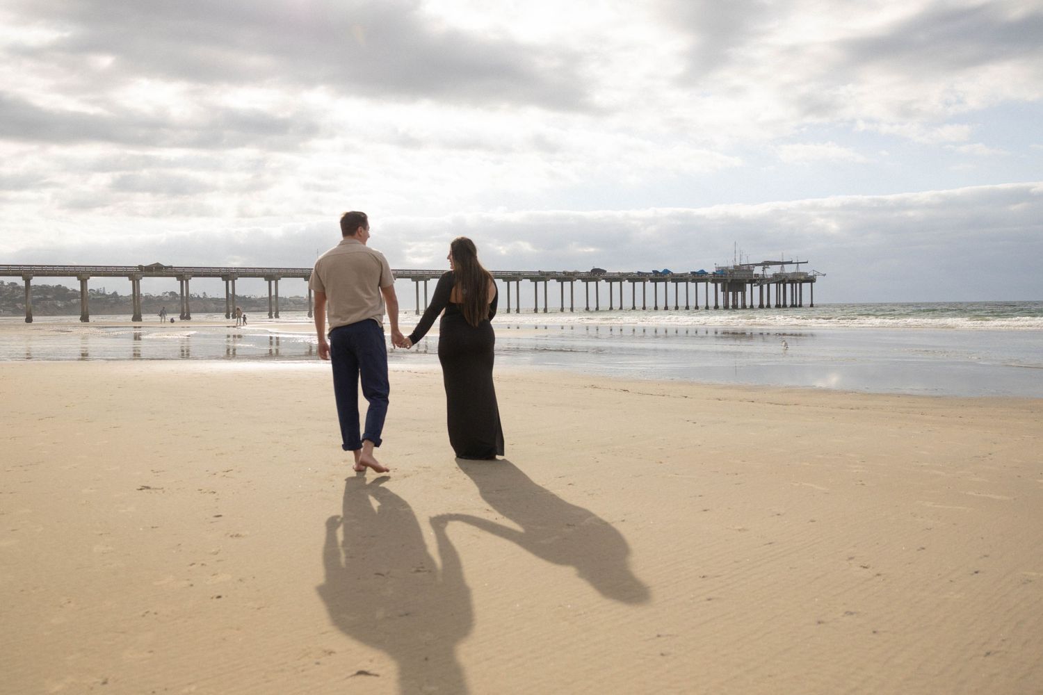 Couple holding hands walking on sandy beach toward ocean pier under cloudy sky. Maternity Photographer San Diego, La Jolla Shores.
