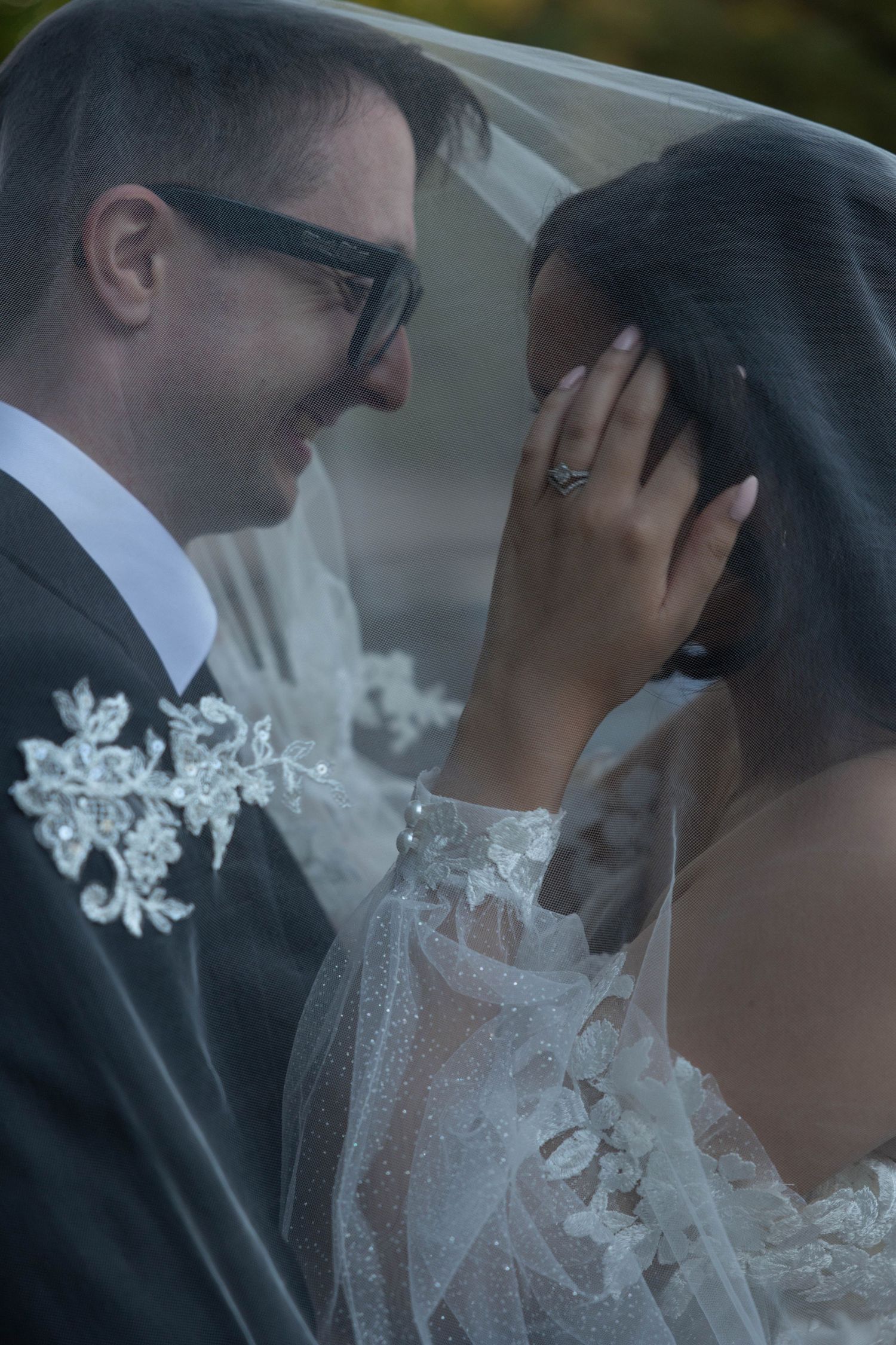 A smiling groom in sunglasses and bride in lace wedding dress share an intimate moment under her veil. Wedding photography.