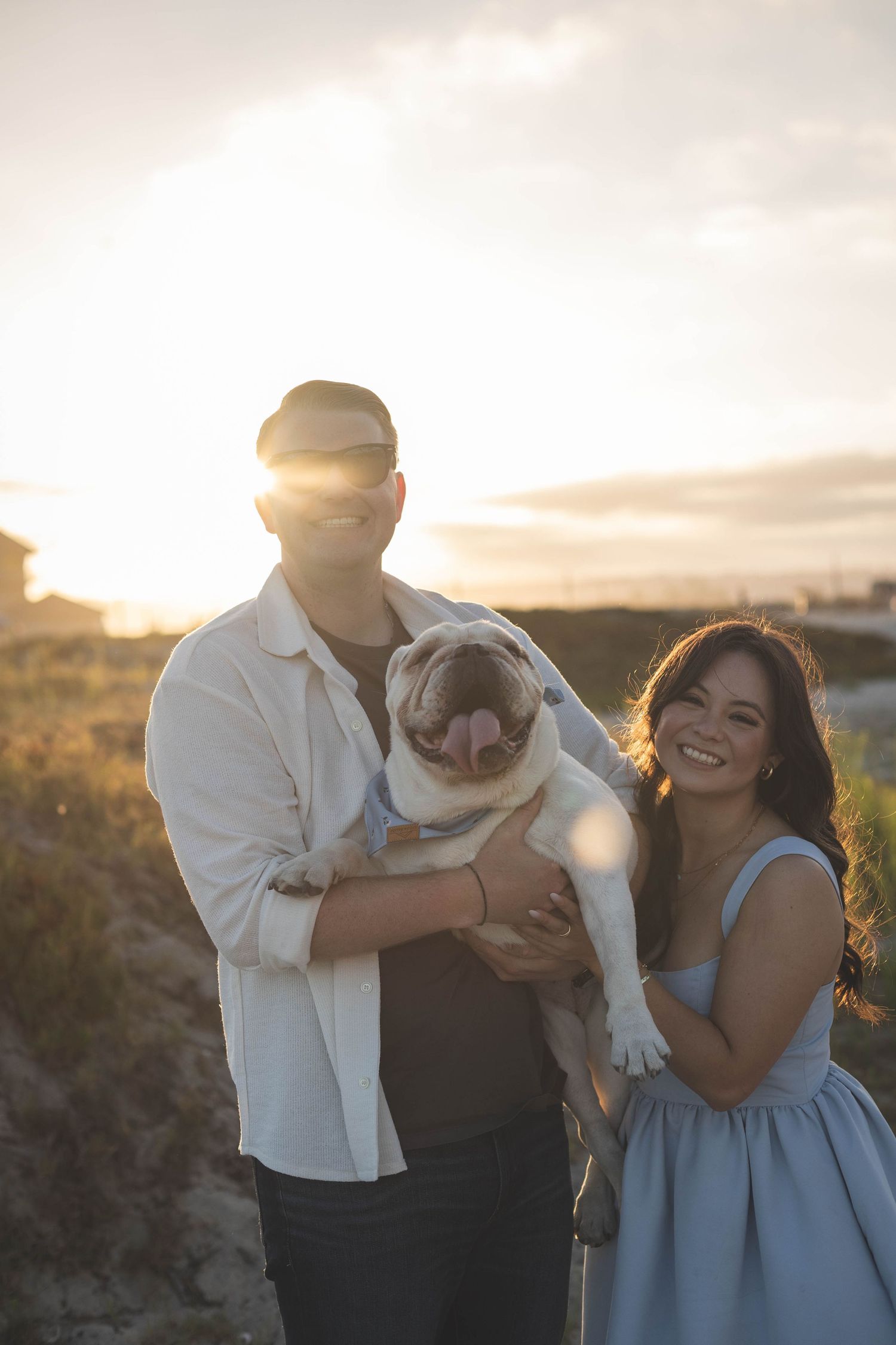 Couple smiling at sunset on beach holding a pug dog, golden hour backlight, woman in blue dress.