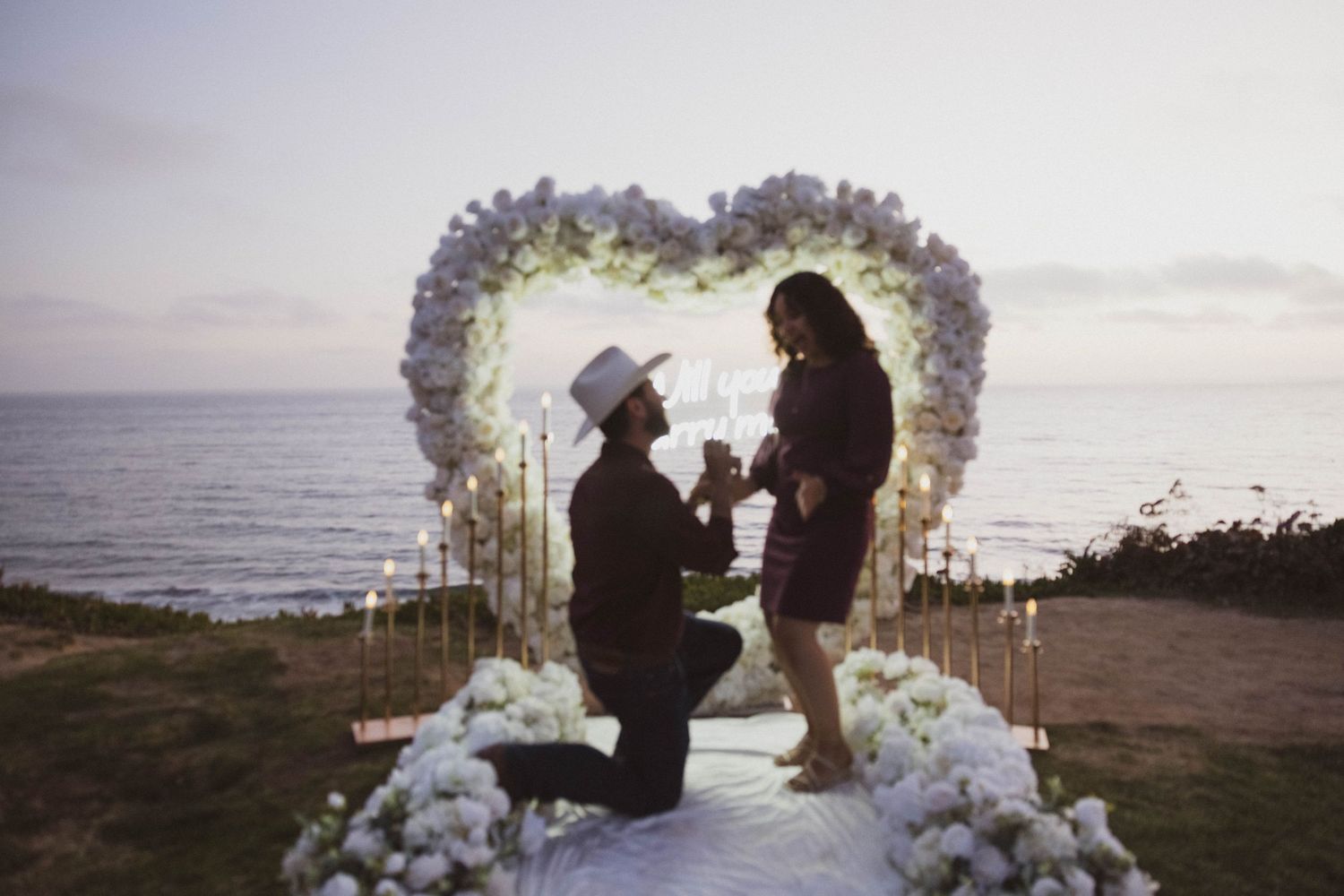 Man proposes to woman on one knee under a floral heart arch with candles by the ocean at sunset. Engagement Photos.