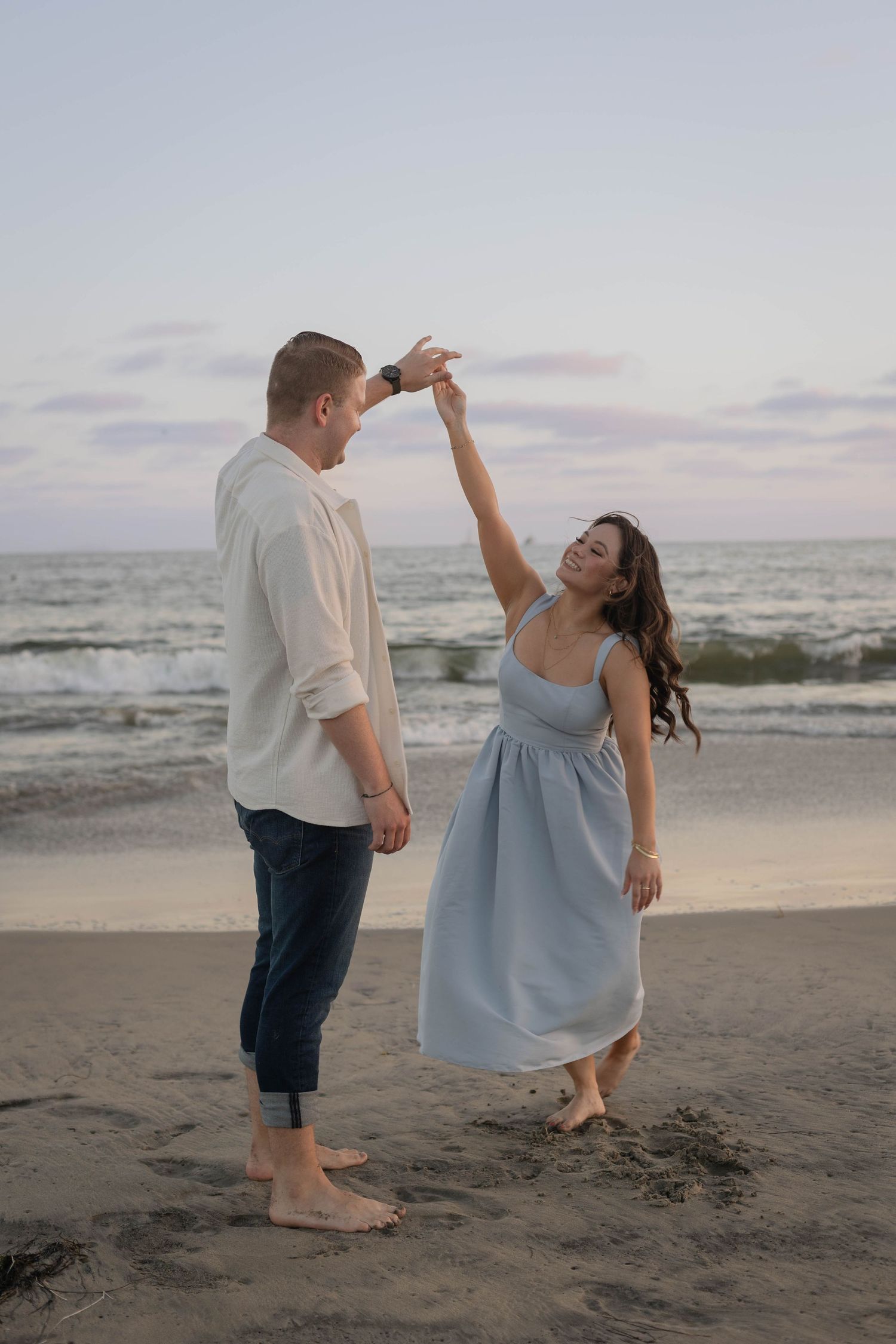 Couple dancing barefoot on a sandy beach at dusk, woman in blue dress twirling under mans raised hand. Coronado beach couples photographer.