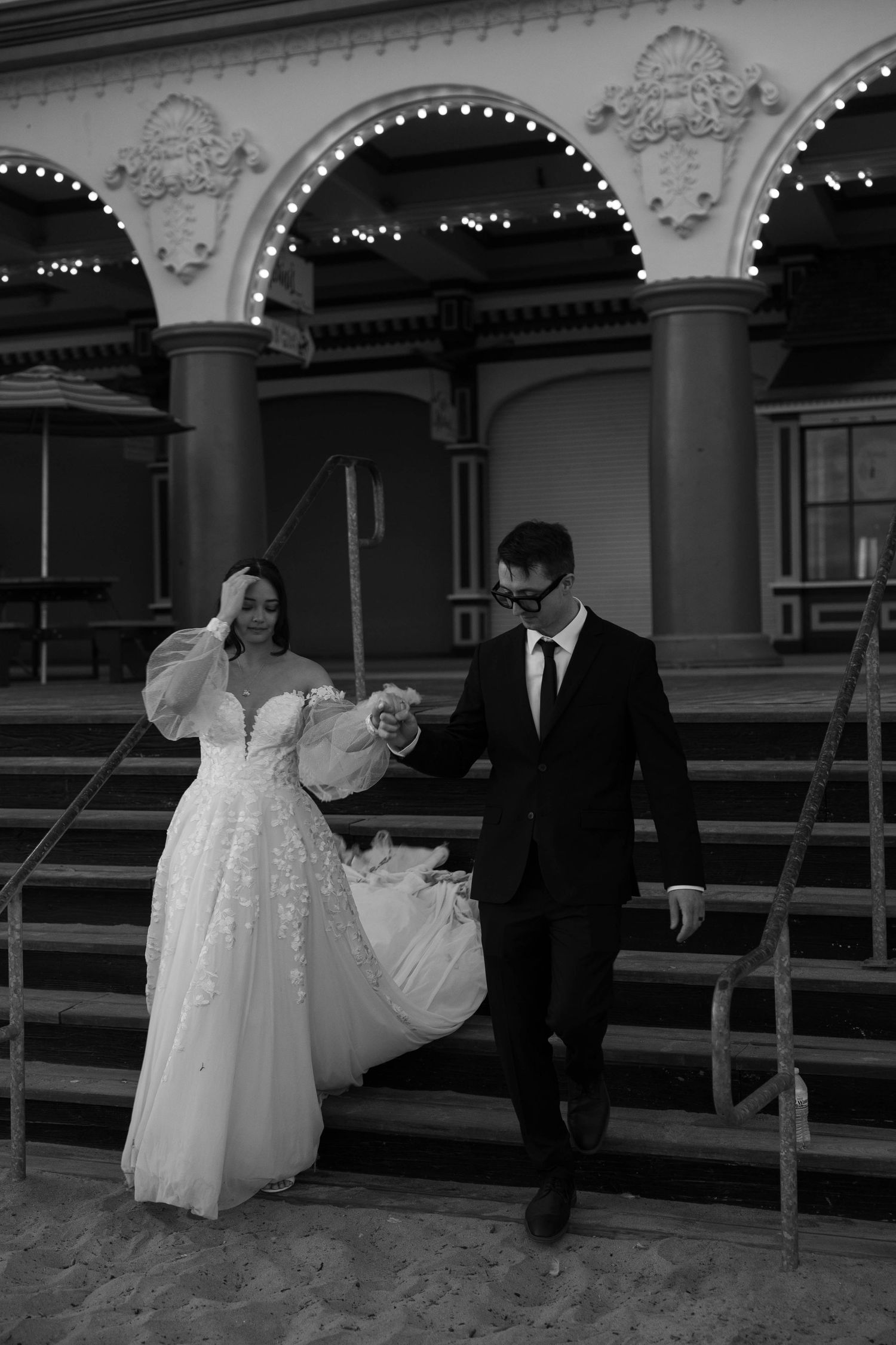 Black and white photo of a bride and groom walking down steps outside an elegant arched building.