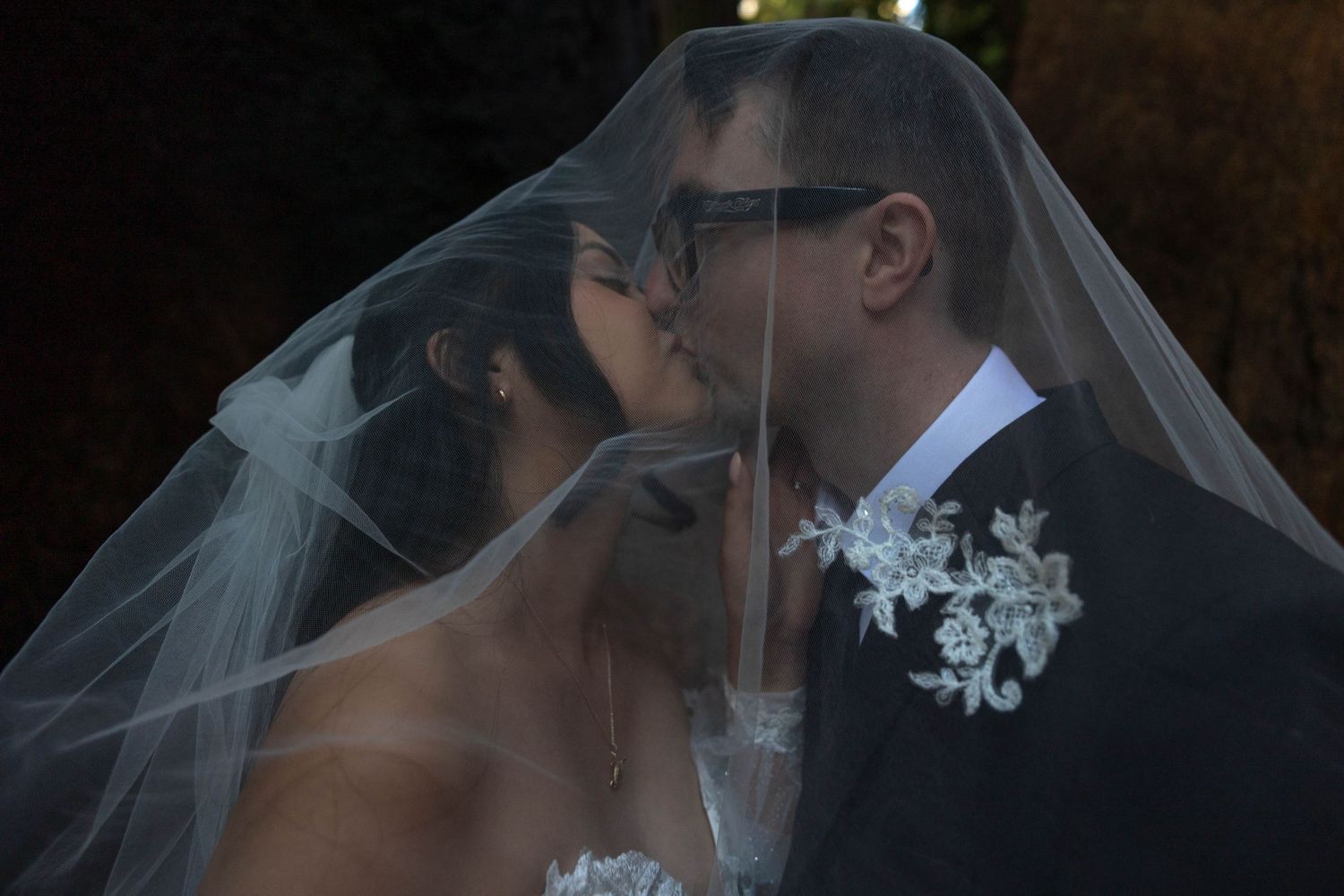 Bride and groom share a romantic kiss beneath the bride's flowing white veil outdoors.