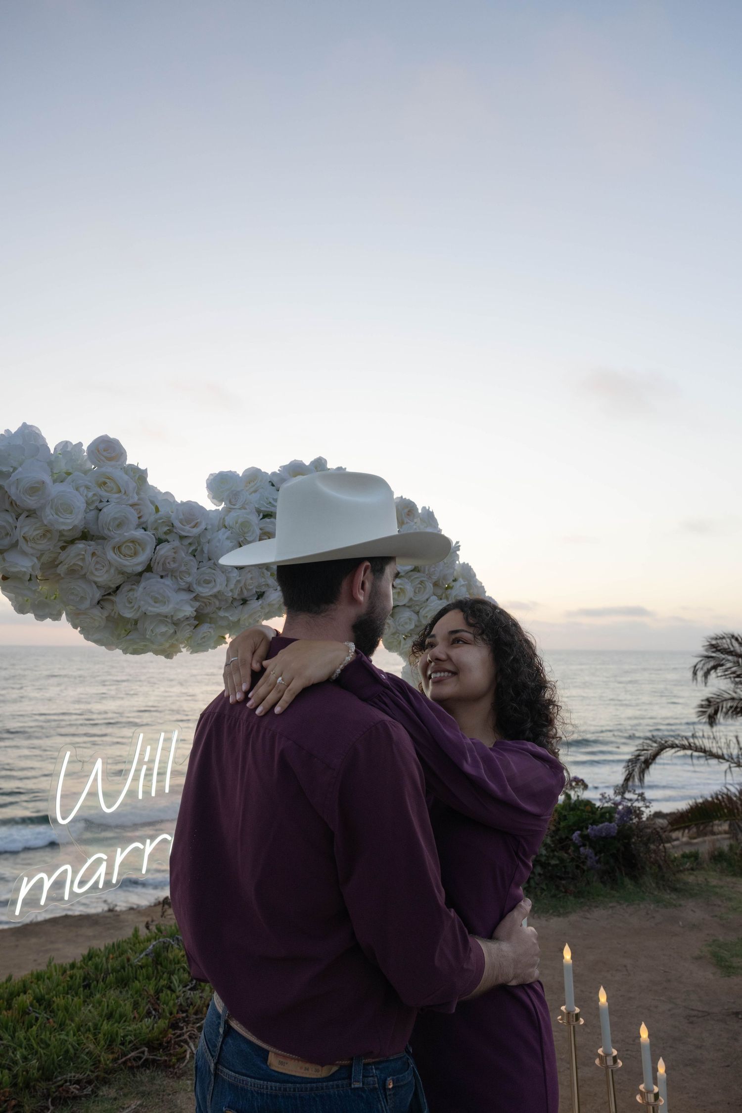 Couple embracing at sunset beach proposal with white floral arch, candles, and will you marry me neon sign. Portrait Photography.