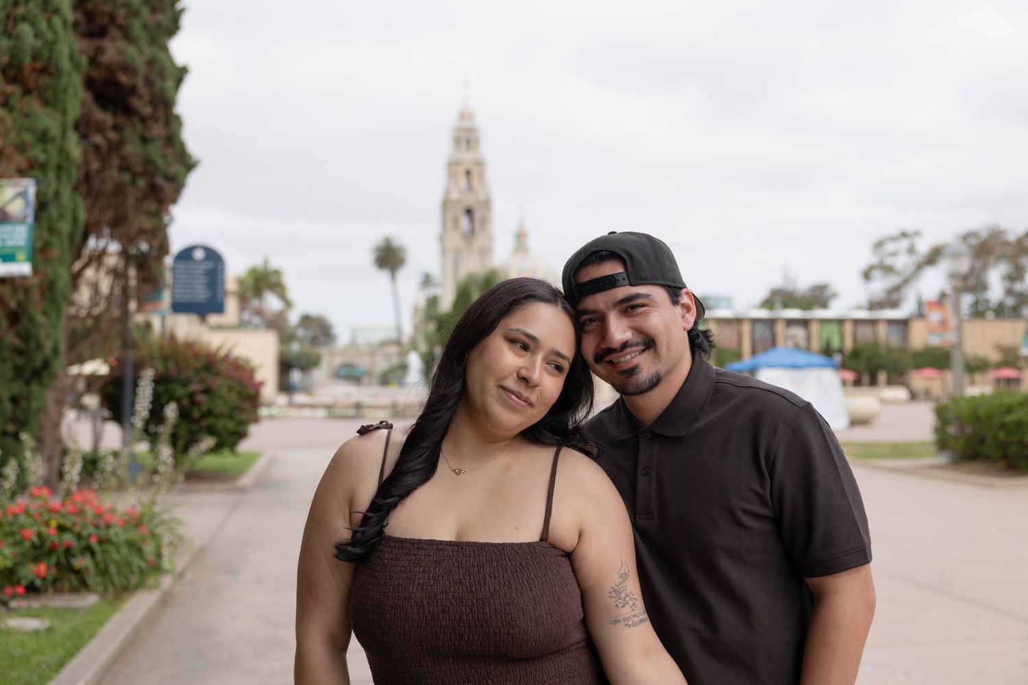A couple poses smiling in front of Balboa Park tower in San Diego, CA on a cloudy day. Portrait photographer.