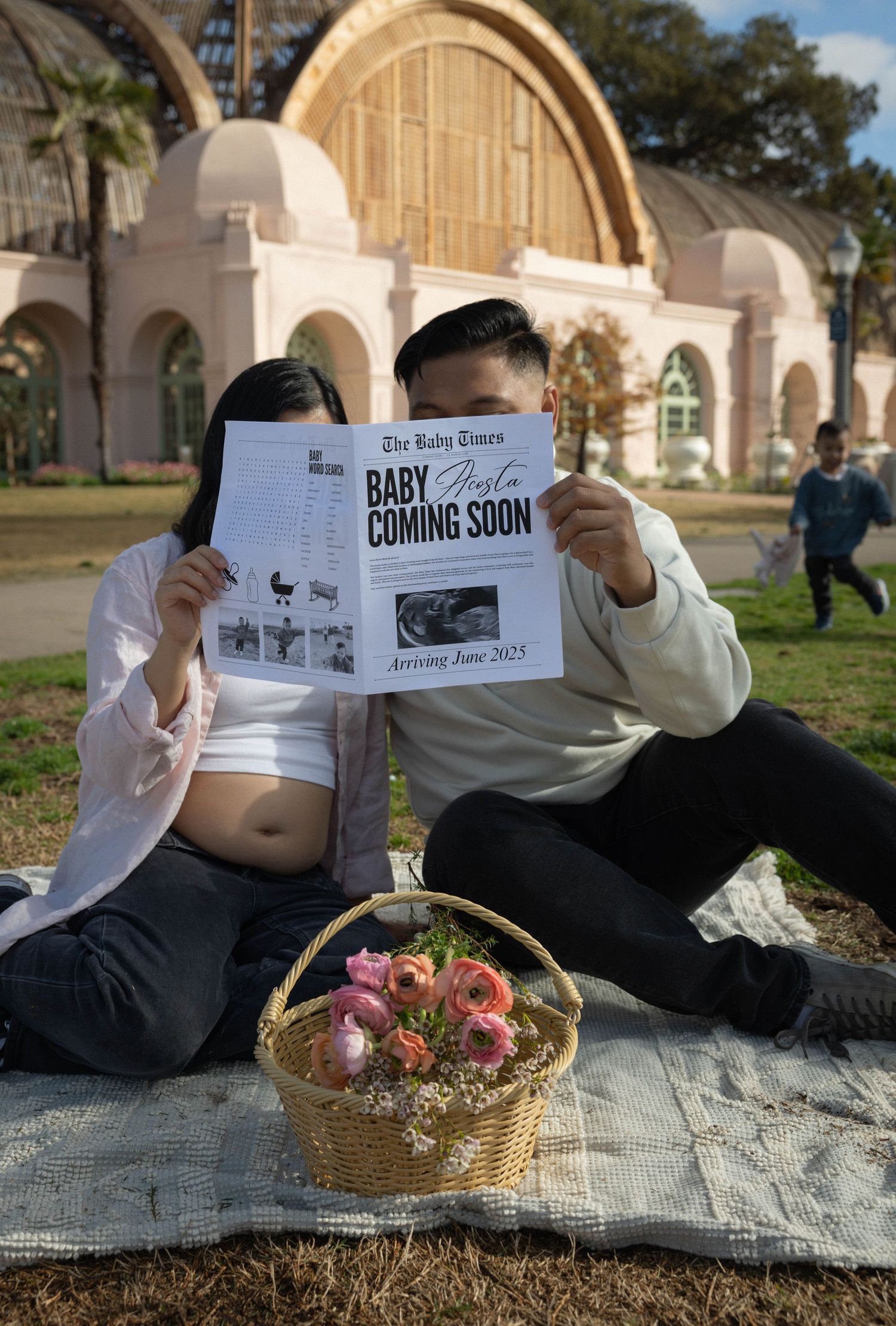 Couple holds pregnancy announcement newspaper at park picnic, revealing baby arriving June 2025, with floral basket. Family photographer SD.