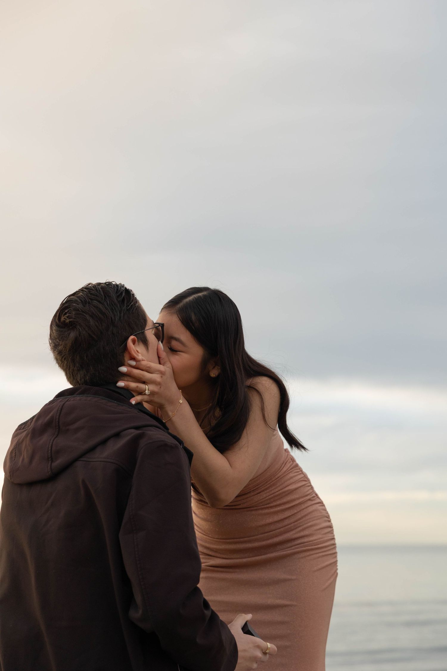 A couple shares a romantic kiss on the beach at dusk, woman in pink dress with engagement ring visible. San Diego Engagement Photographer.