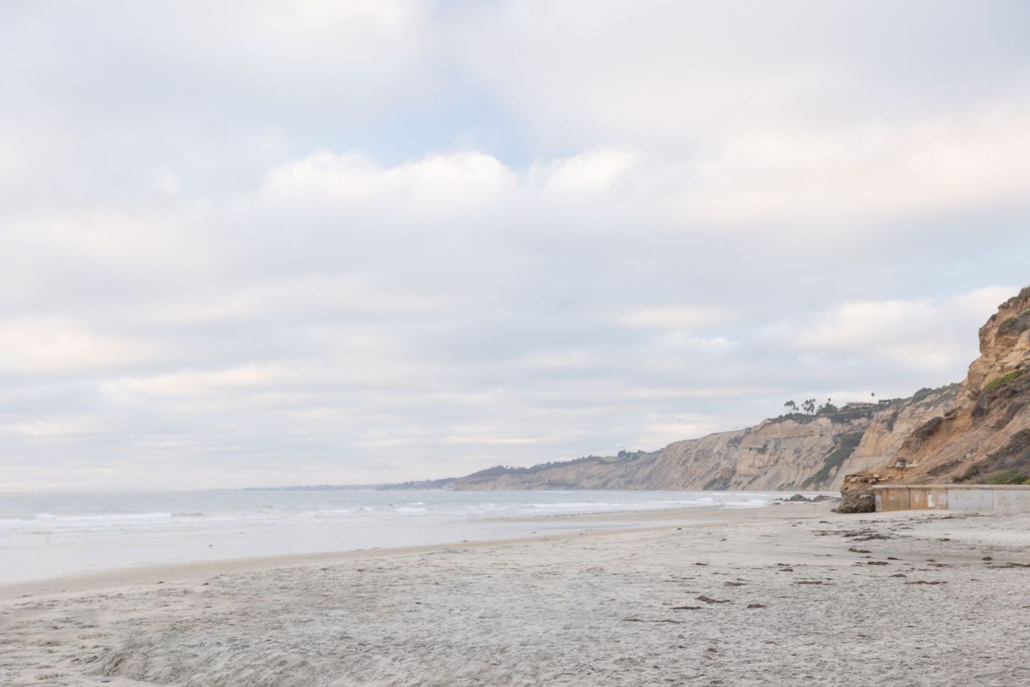 A serene coastal beach scene with sandy shores, gentle waves, rocky cliffs, and a cloudy sky in the background. La Jolla Shores, San Diego.