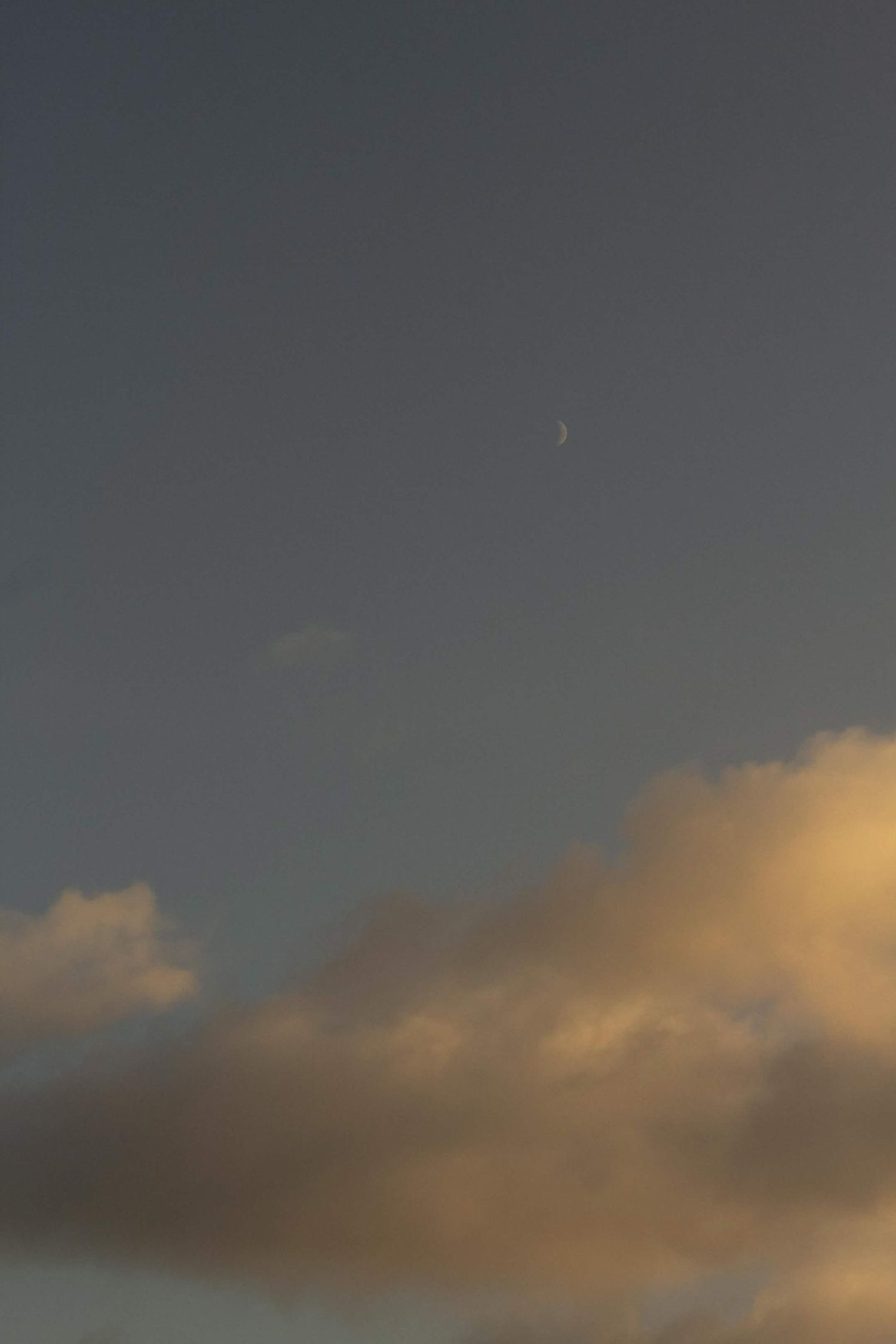 Serene twilight sky with a crescent moon visible above warm golden-toned clouds at dusk.