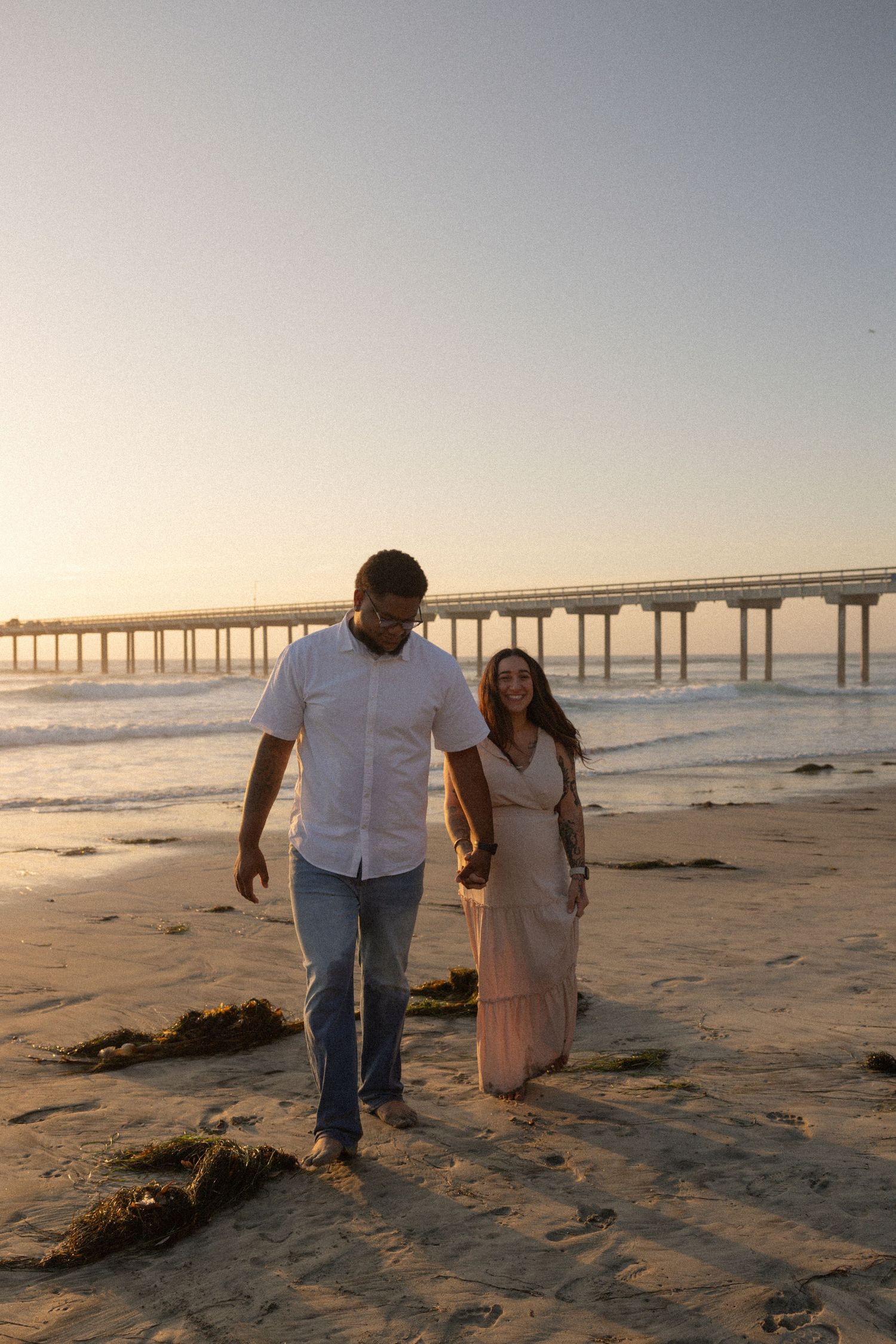 Couple walking hand in hand on a sandy beach at sunset with a pier in the background. Maternity Photographer San Diego California. Golden.