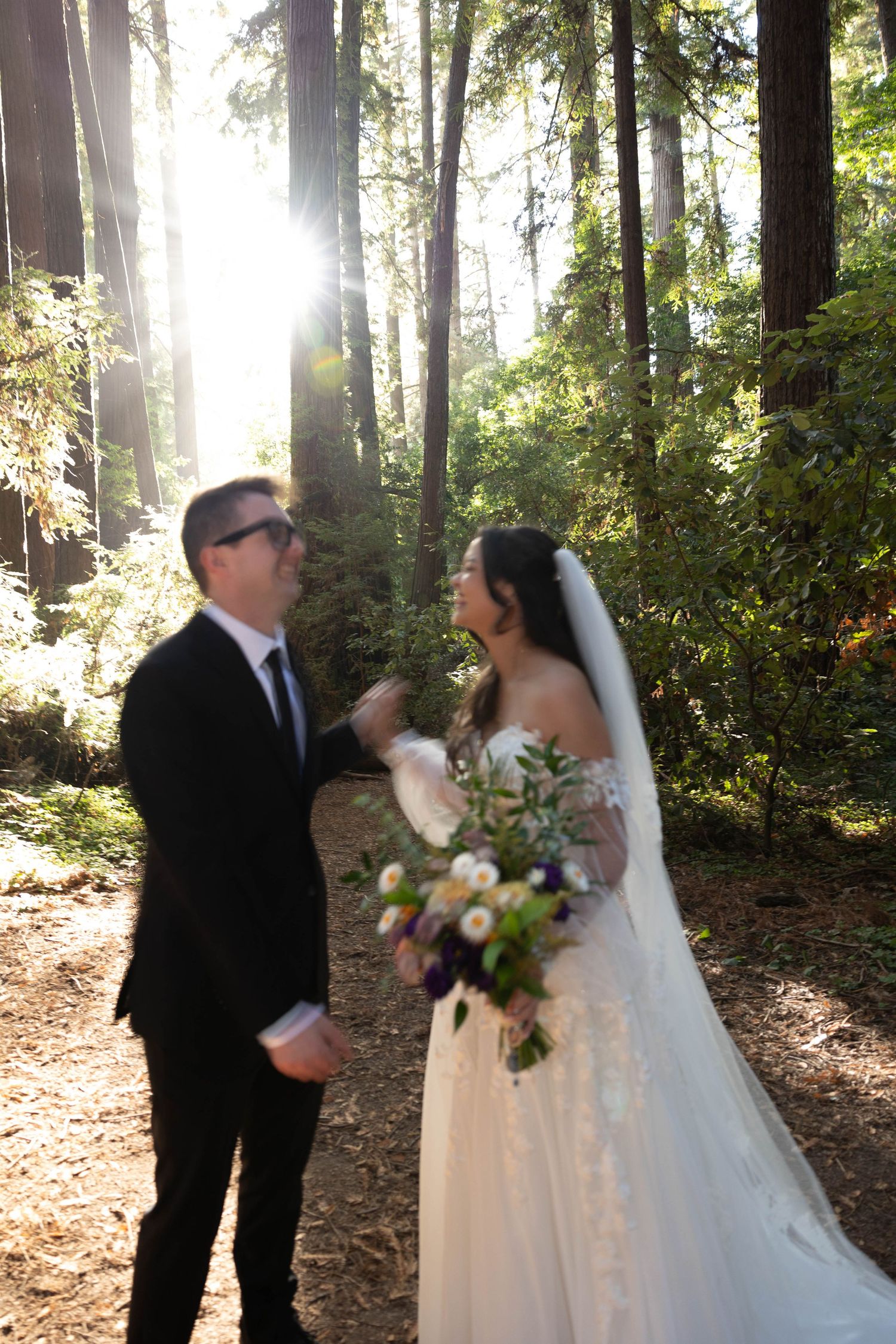 Couple laughs joyfully together in a redwood forest, bride in lace gown holding bouquet with sunlight streaming through trees.