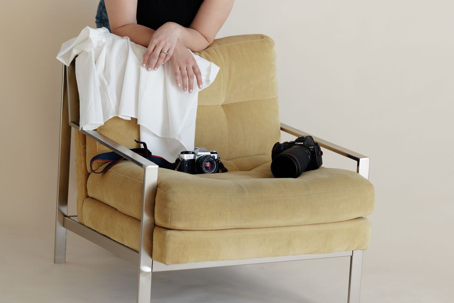 Person standing behind a yellow velvet modern armchair, on the cough is a draped white shirt and two cameras.