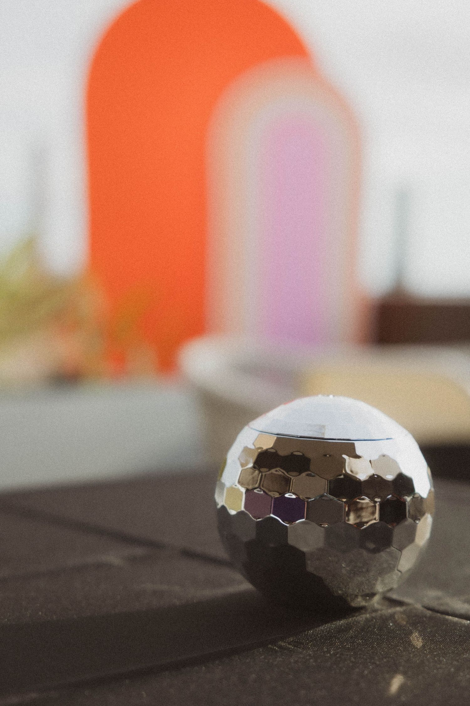 Small silver mirrored disco ball in sharp focus against blurred colorful arch decorations on a dark surface.
