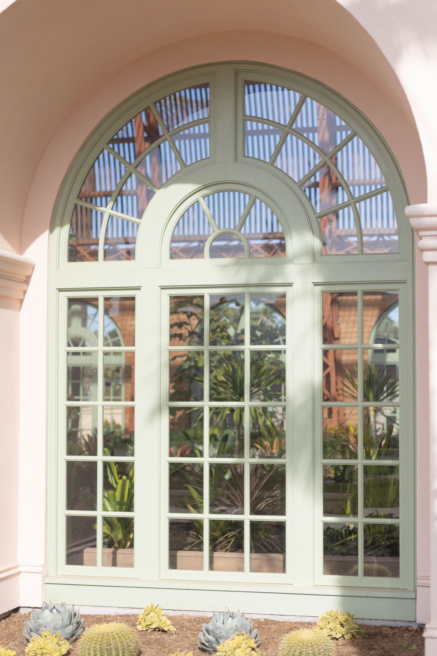 Elegant arched green window with grid panes on a pink stucco building, surrounded by cacti and tropical plants.