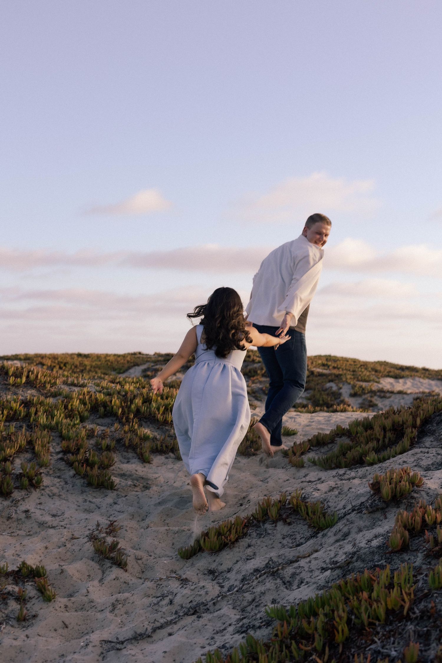 Couple holding hands running barefoot on sandy beach dunes at sunset, woman in white dress, man in white shirt. Coronado Beach, San Diego.