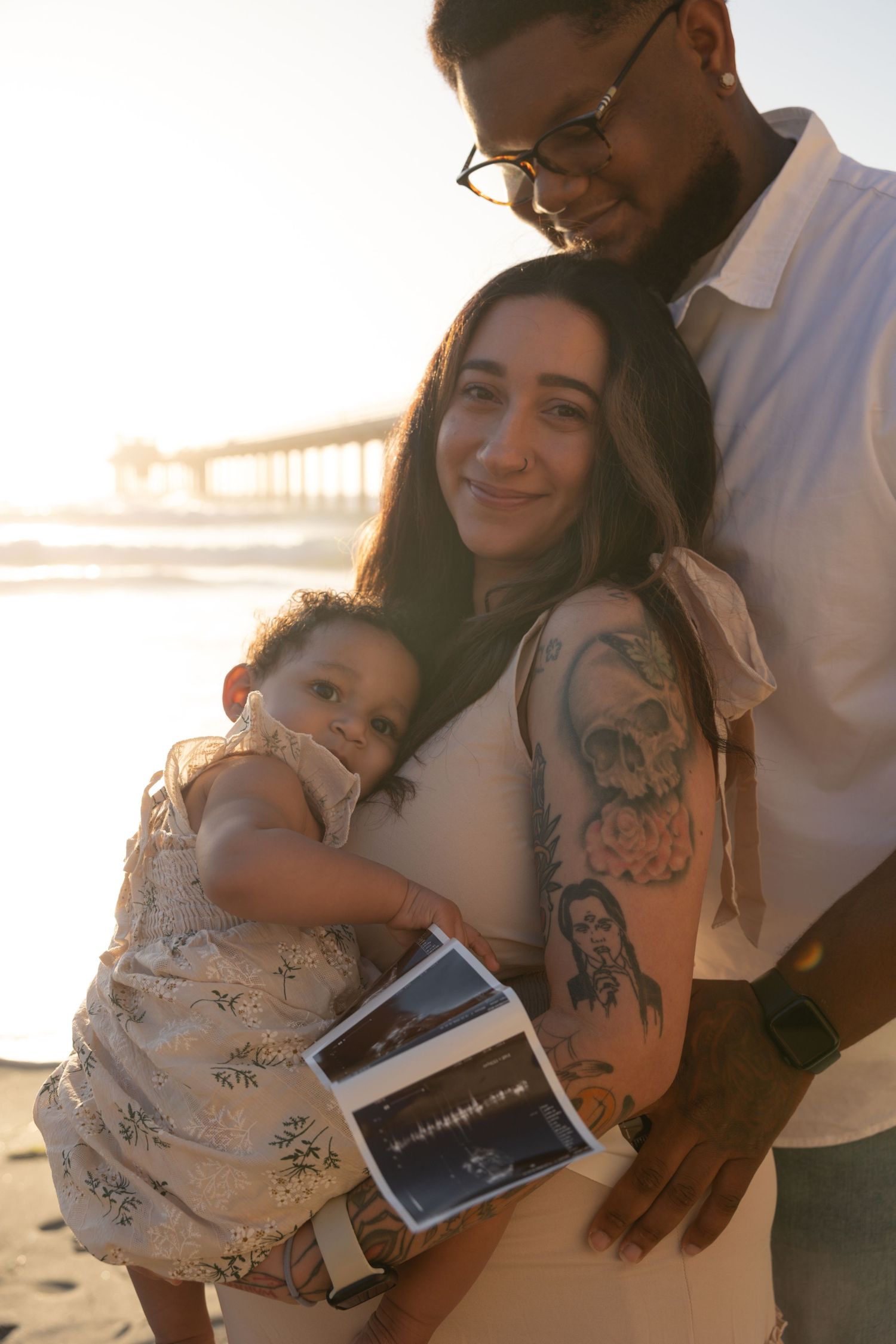 A family poses on a beach at sunset, holding a child and ultrasound photos. Maternity Photographer San Diego California. Golden Hour.
