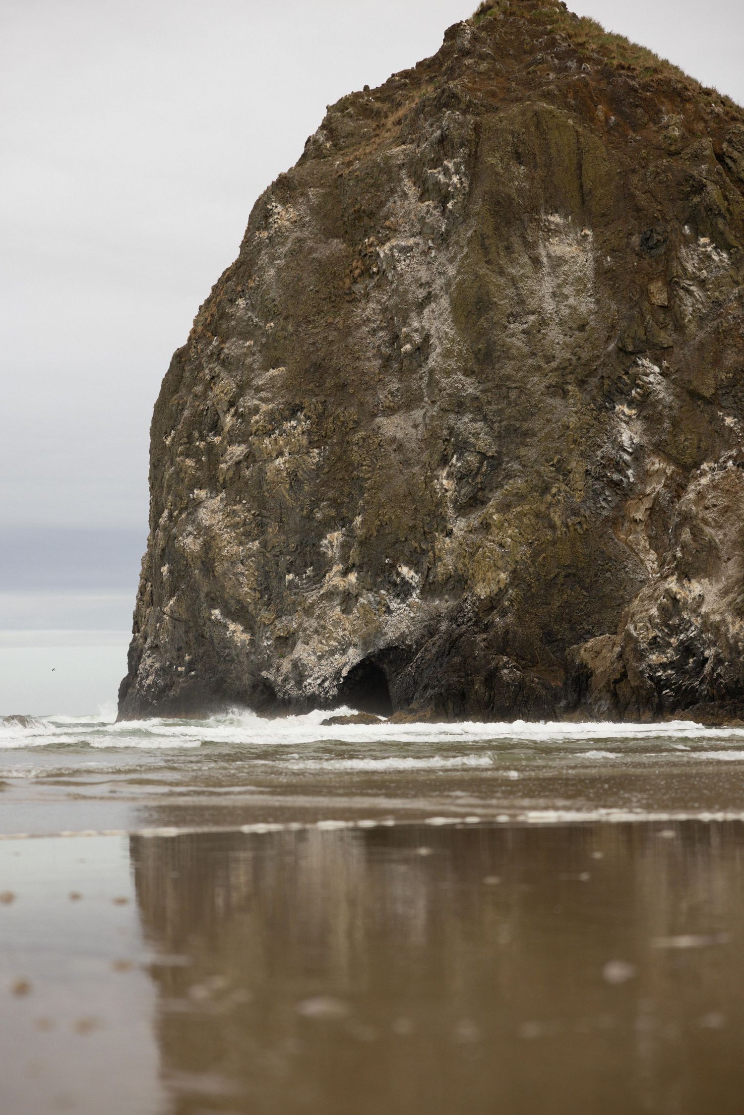 Massive Haystack Rock at Cannon Beach, Oregon, with ocean waves and wet sand reflection on an overcast day.