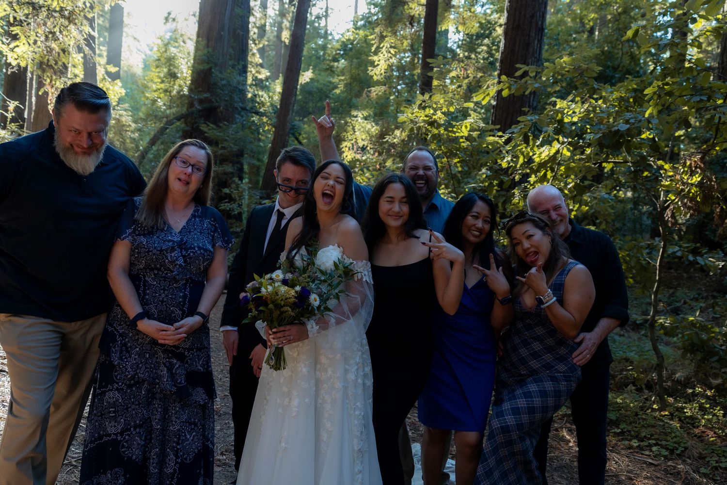 Wedding party celebrating in a sunlit redwood forest, bride in white gown holding bouquet with joyful guests. Wedding photography.