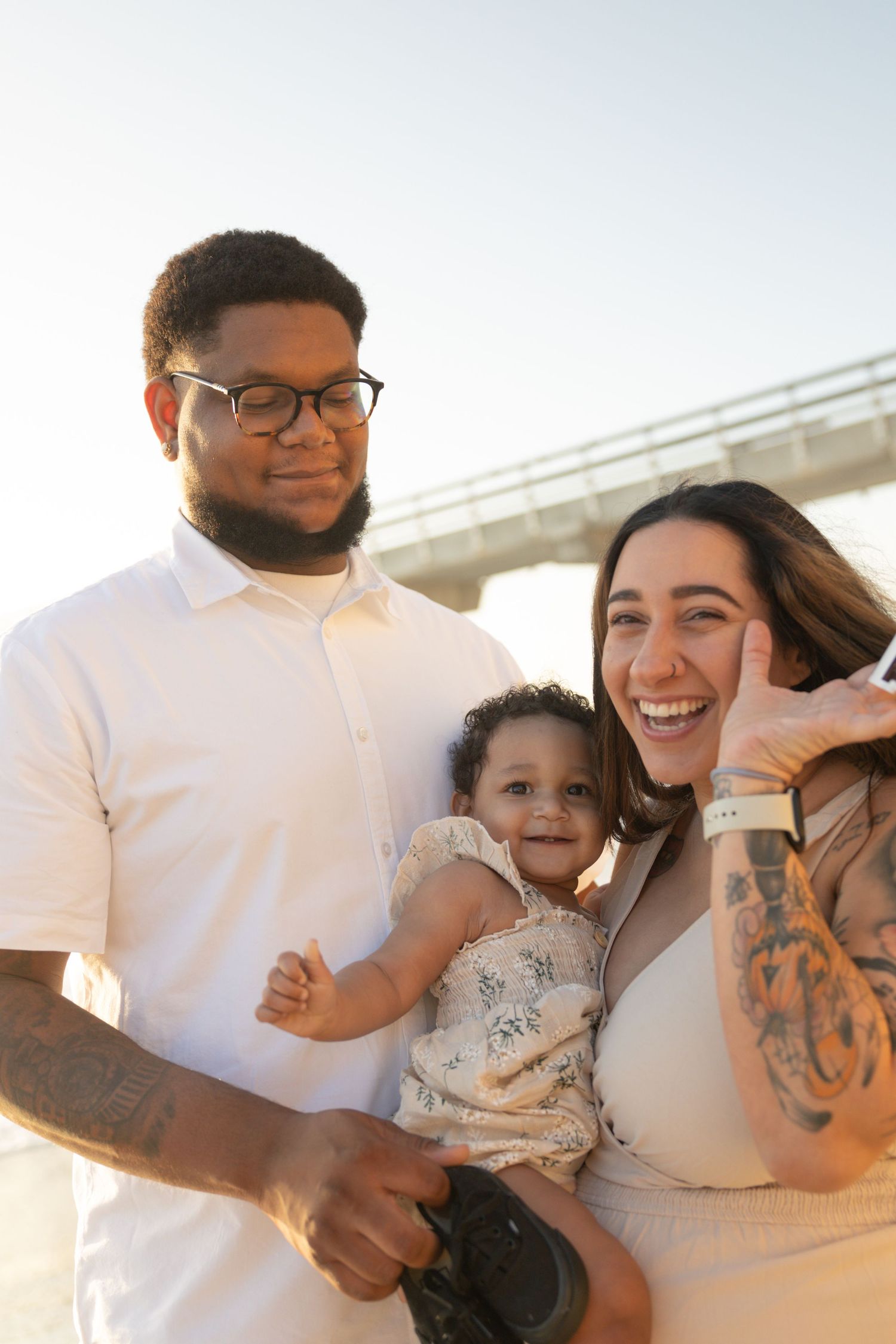 Happy family smiling outdoors near a bridge, parents holding their baby girl in a floral outfit at sunset. Maternity Photographer San Diego