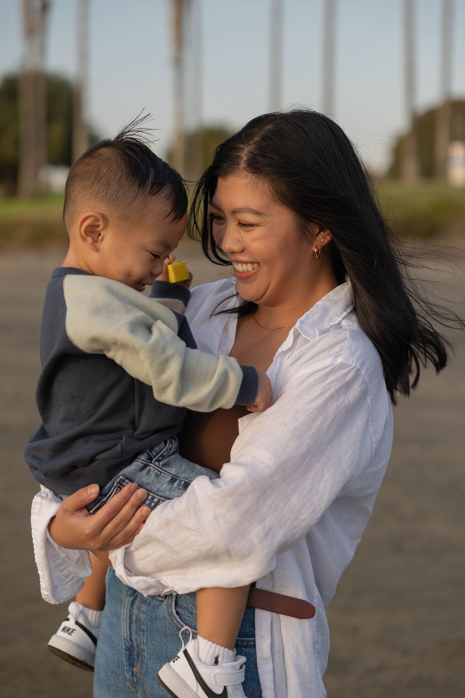 A smiling woman in a white shirt holds a young toddler boy outdoors near palm trees at golden hour. Family Photographer.