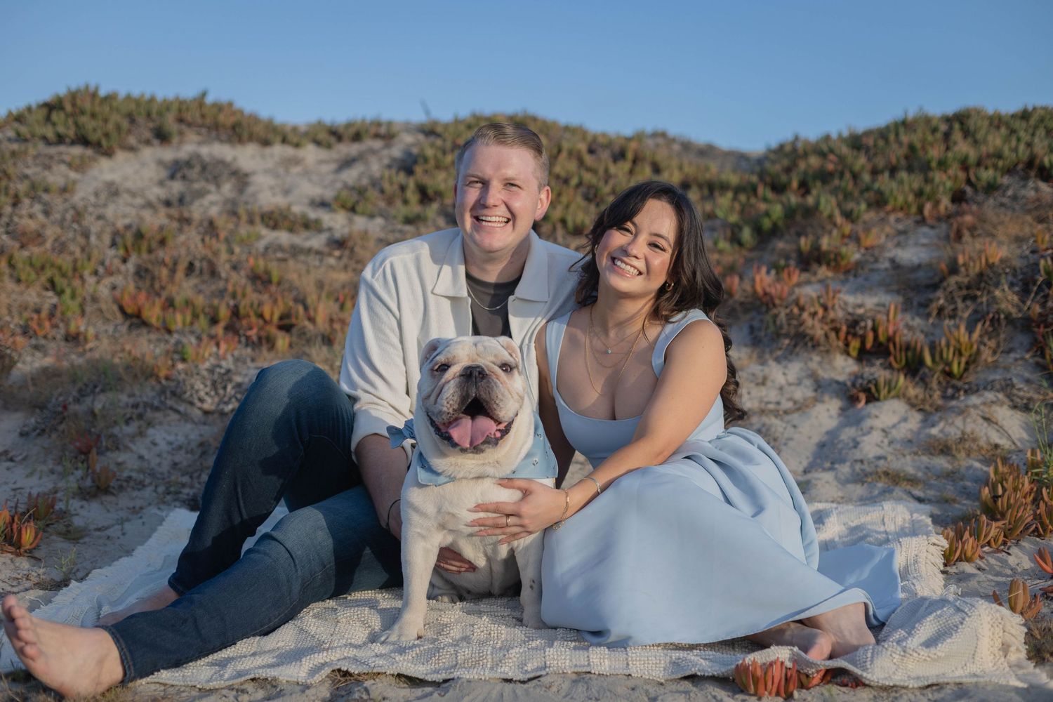 Smiling couple sitting on beach sand with their English Bulldog, sandy dunes and blue sky behind them.
