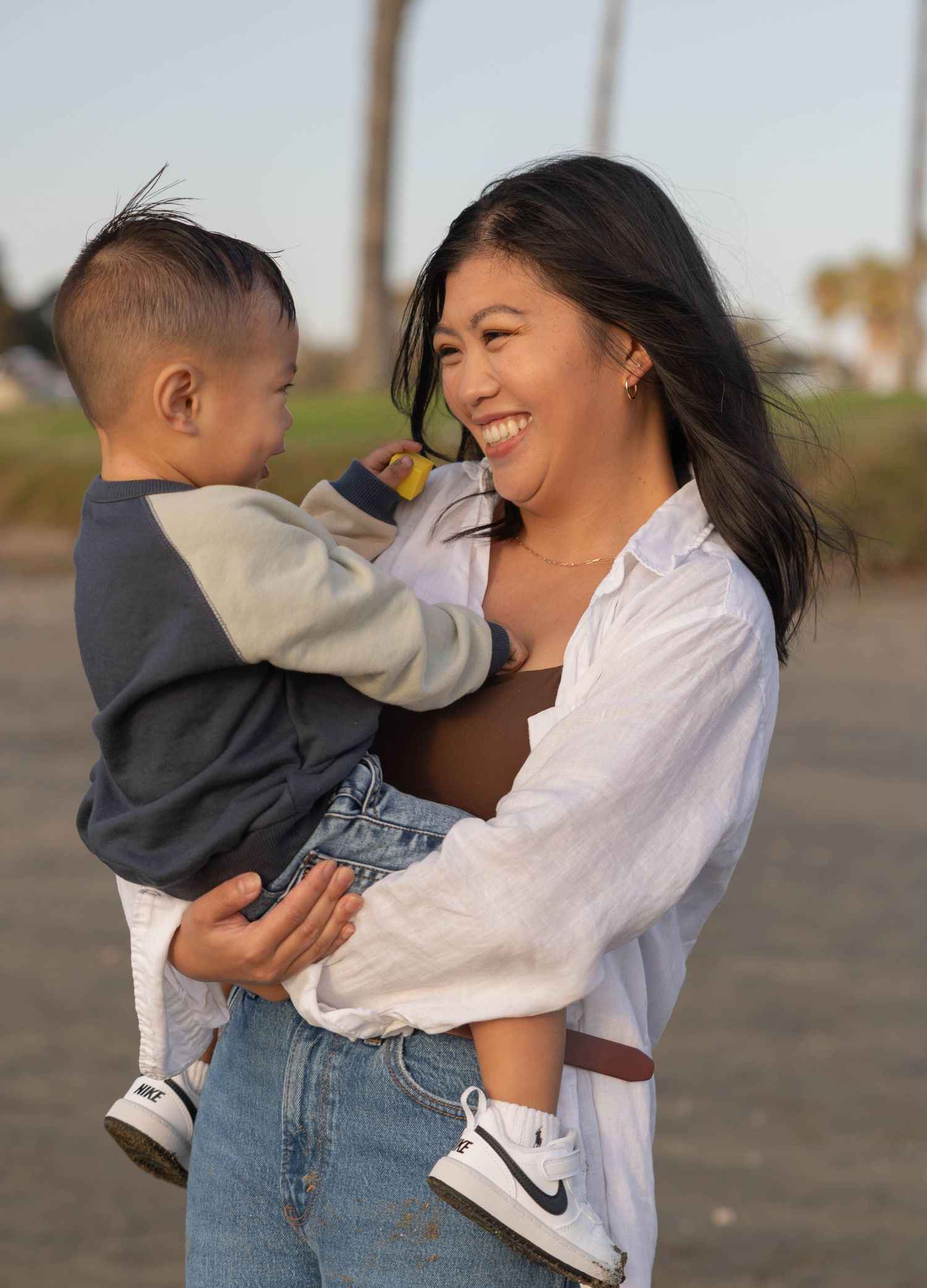 A smiling woman holds a young toddler outdoors, both wearing casual clothes, sharing a joyful moment together. Family Photographer.