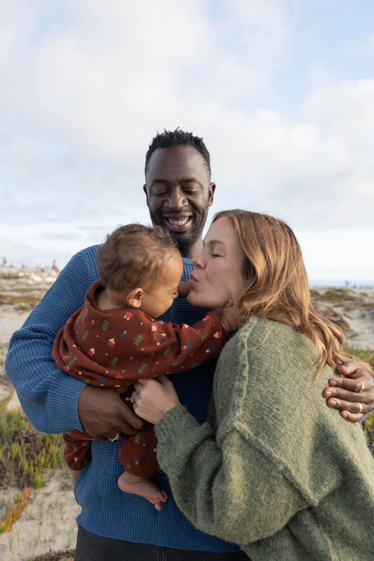 Happy family with mother kissing baby held by smiling father at a sunny beach. Family Photographer.