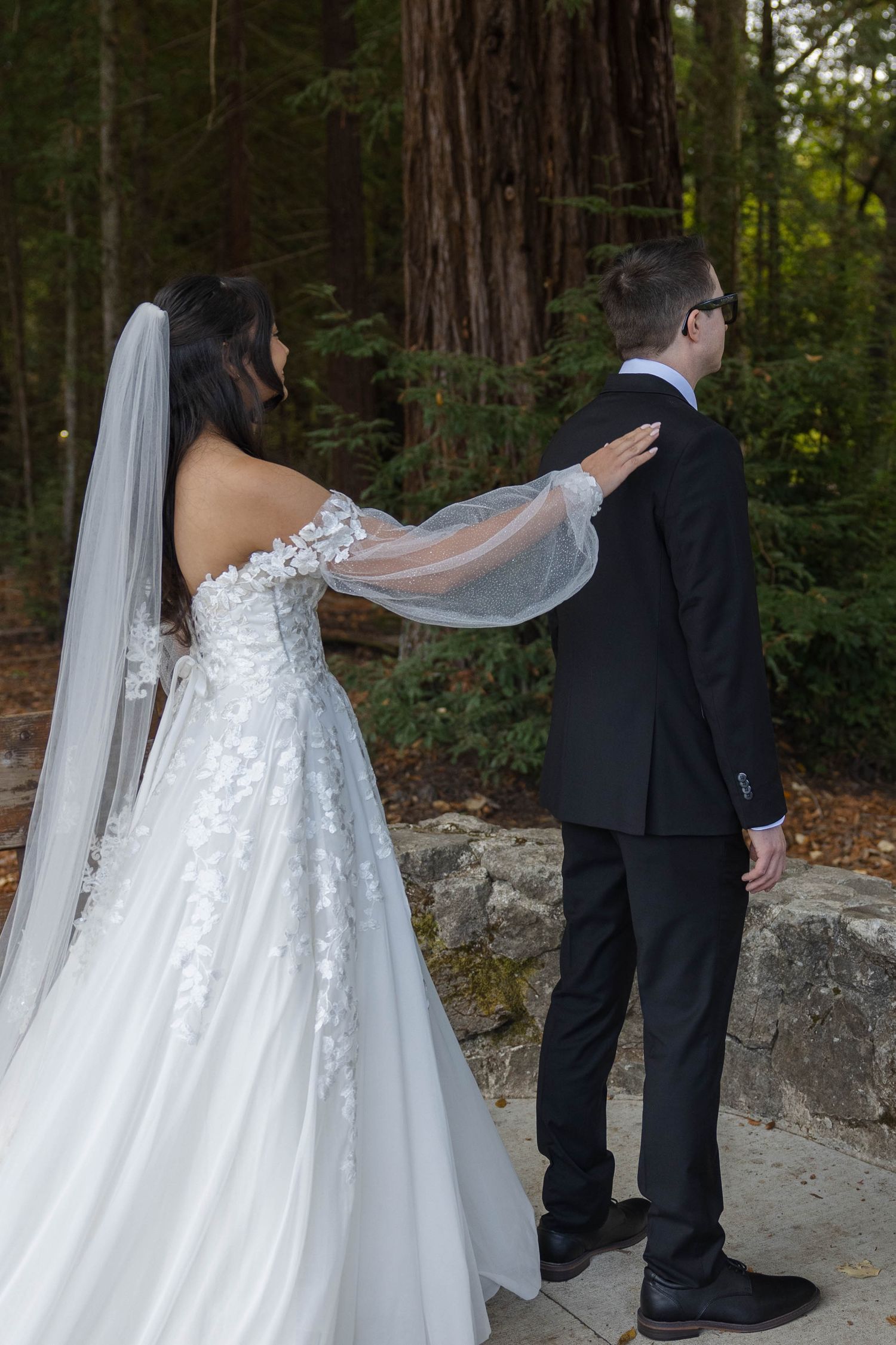 Bride in lace gown touches groom's shoulder during first look in redwood forest.