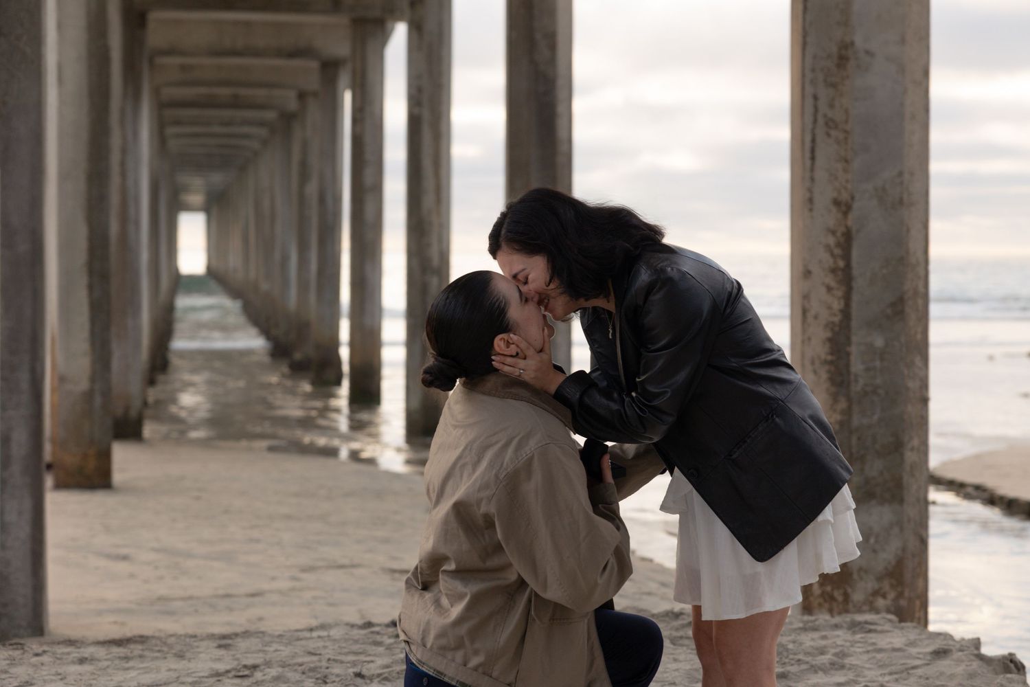 Couple sharing a romantic kiss under a beach pier during a marriage proposal on a cloudy day. Proposal Photos. Engagement Photography.