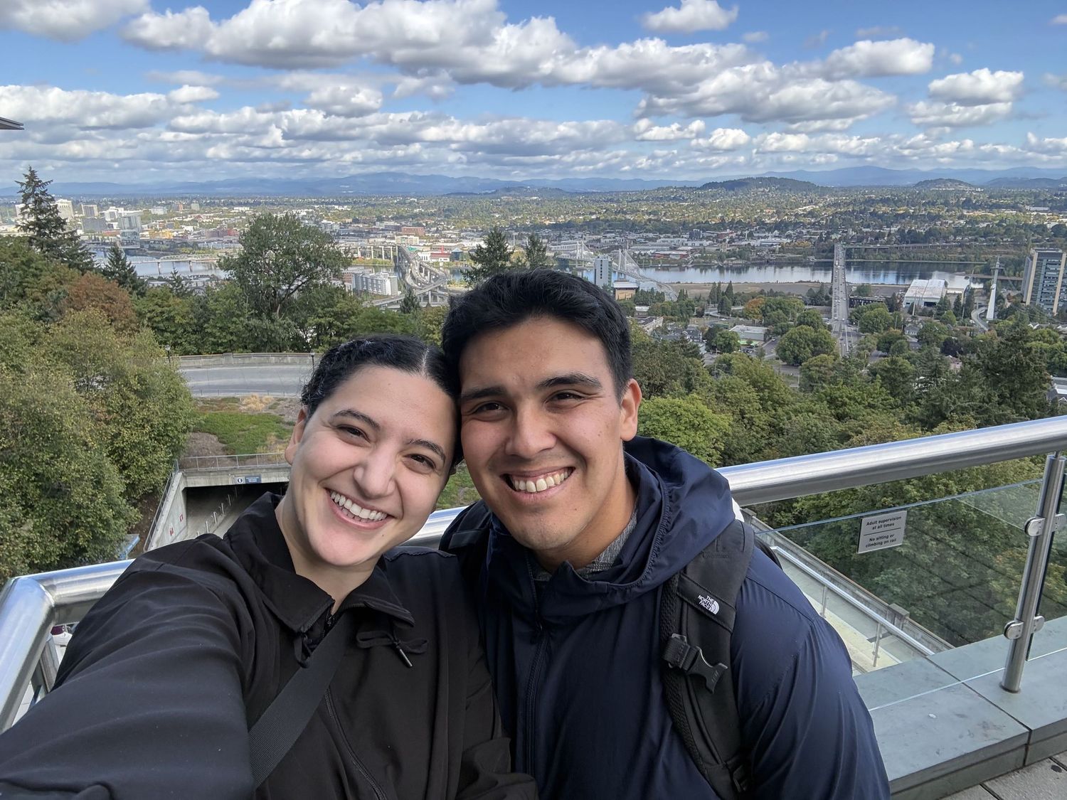 Two smiling people take a selfie on a scenic overlook with a panoramic city and river view on a cloudy day.