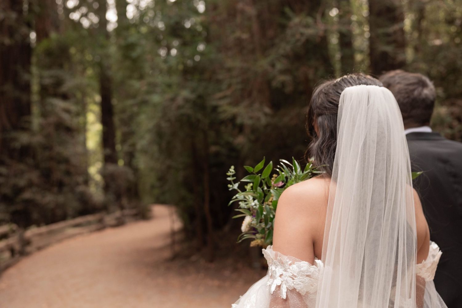 Bride and groom walking through a redwood forest path, bride wearing lace gown, veil, and greenery bouquet. Wedding photographer.