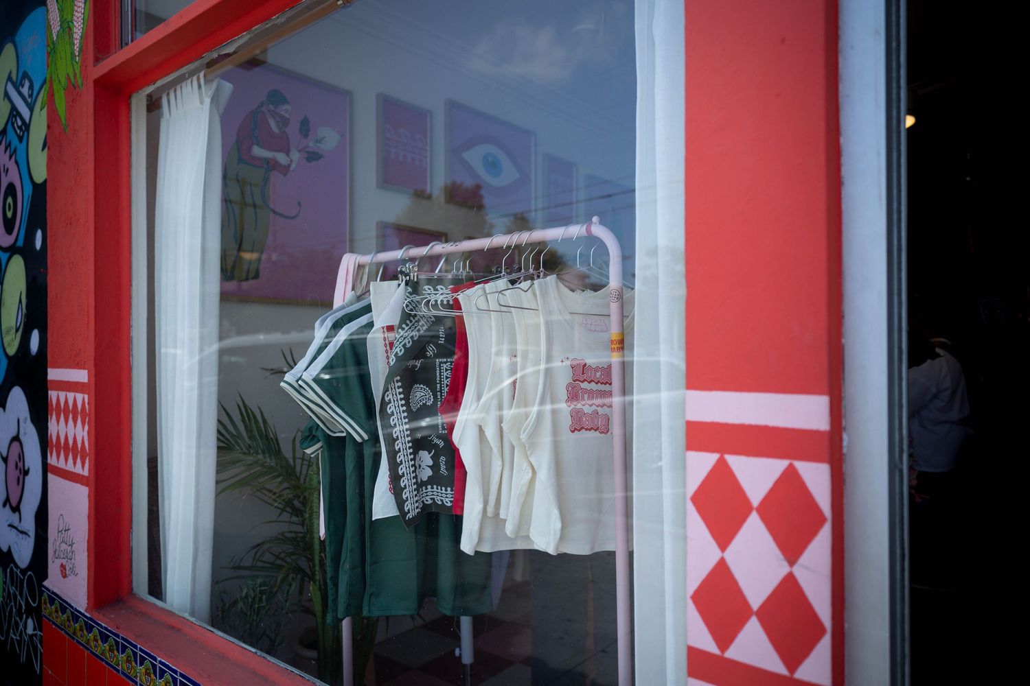 Colorful boutique shop window display with clothing on hangers including green and white shirts on red-framed window.